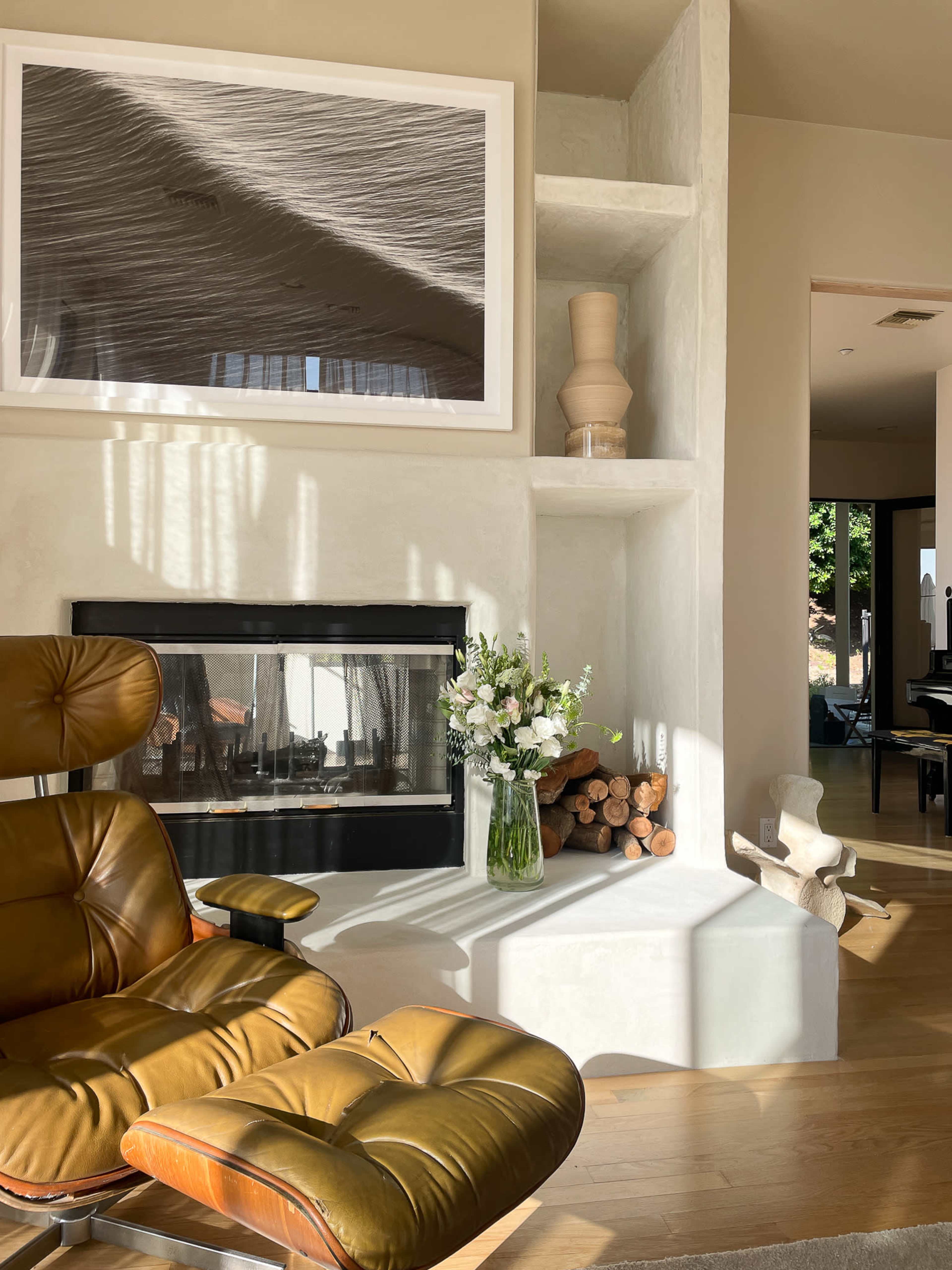 A mid-century modern chair is positioned near a fireplace, which is adorned with a vase of flowers and stacked logs, all bathed in sunlight streaming through a nearby window.