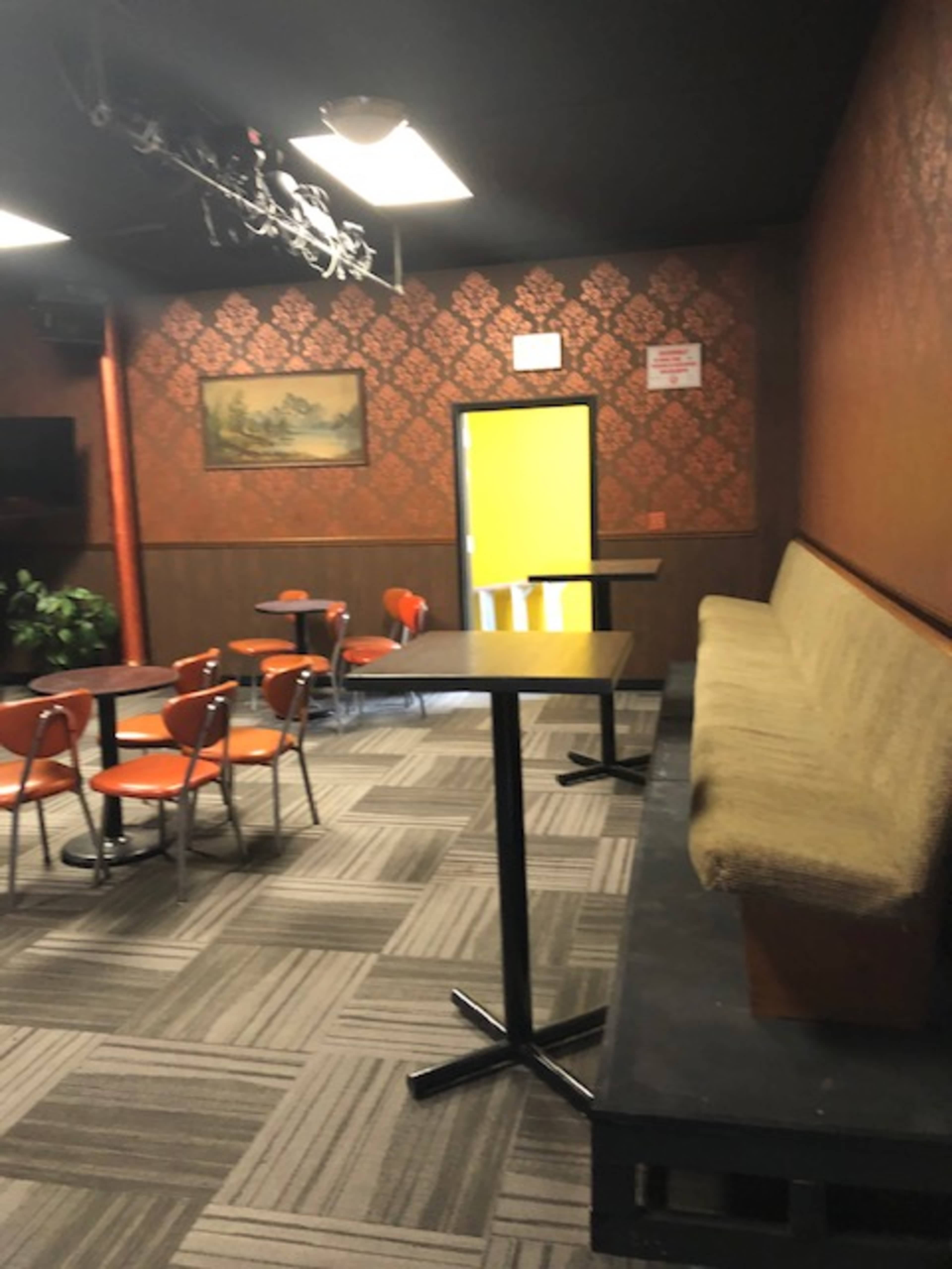 The image shows a small room with orange chairs and tables, a large bench against the wall, and a bright yellow door at the back.