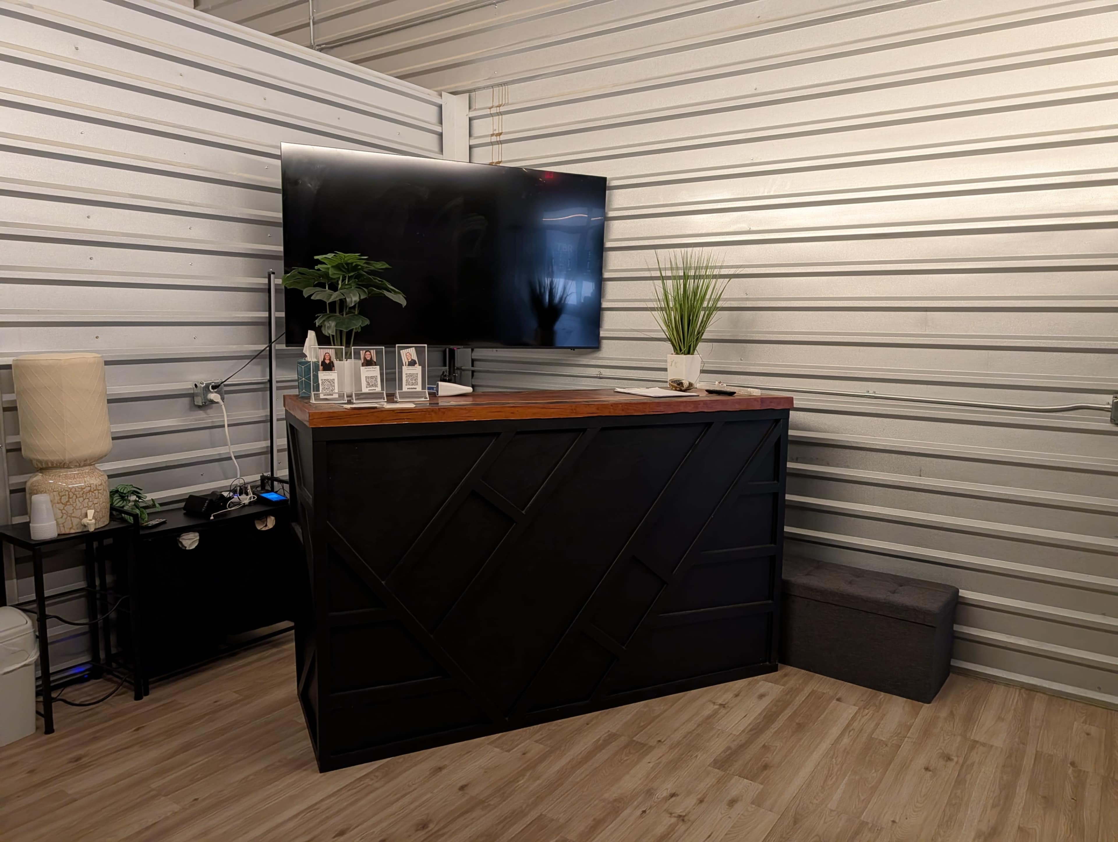 The image shows a reception area with a dark-colored desk, a flat-screen TV mounted on the wall, a small plant, and a lamp beside it, all set against a backdrop of metallic storage unit walls.
