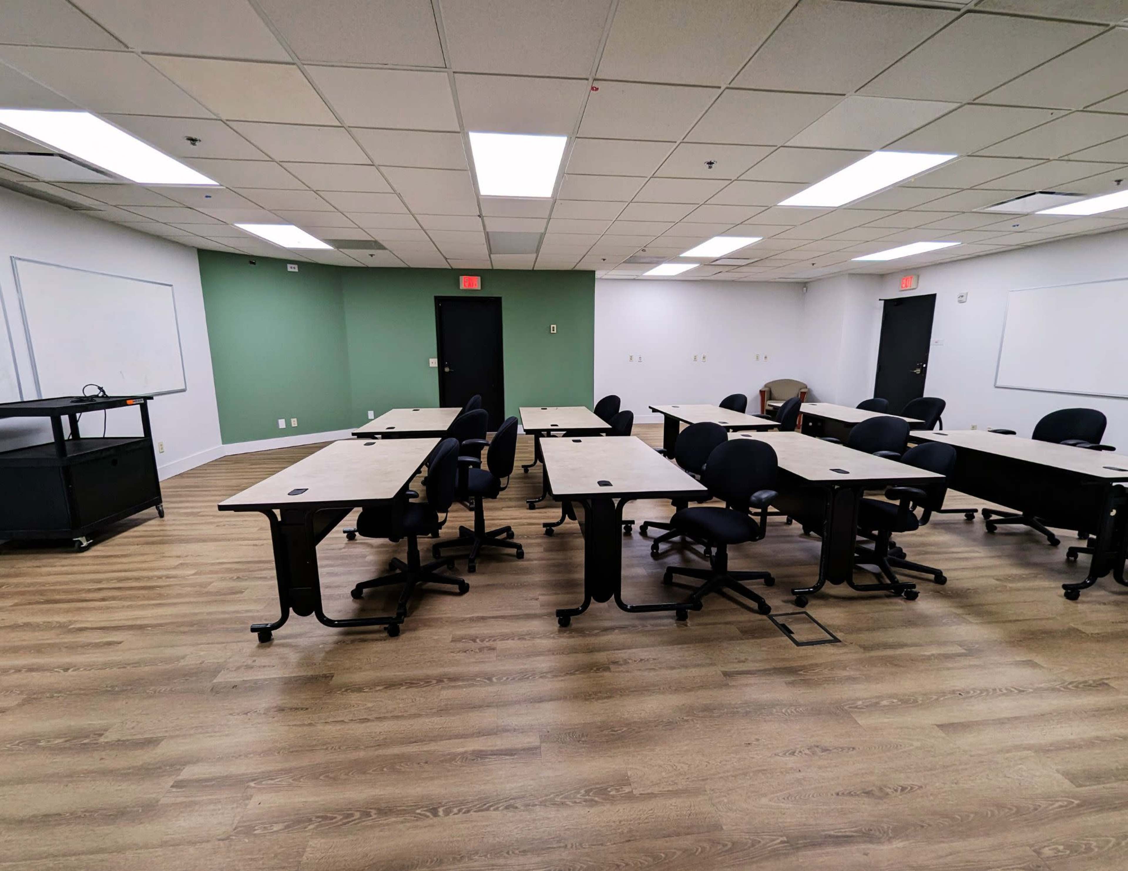 A classroom with multiple desks arranged in a U-shape, surrounded by green and white walls and equipped with a black presentation cart.
