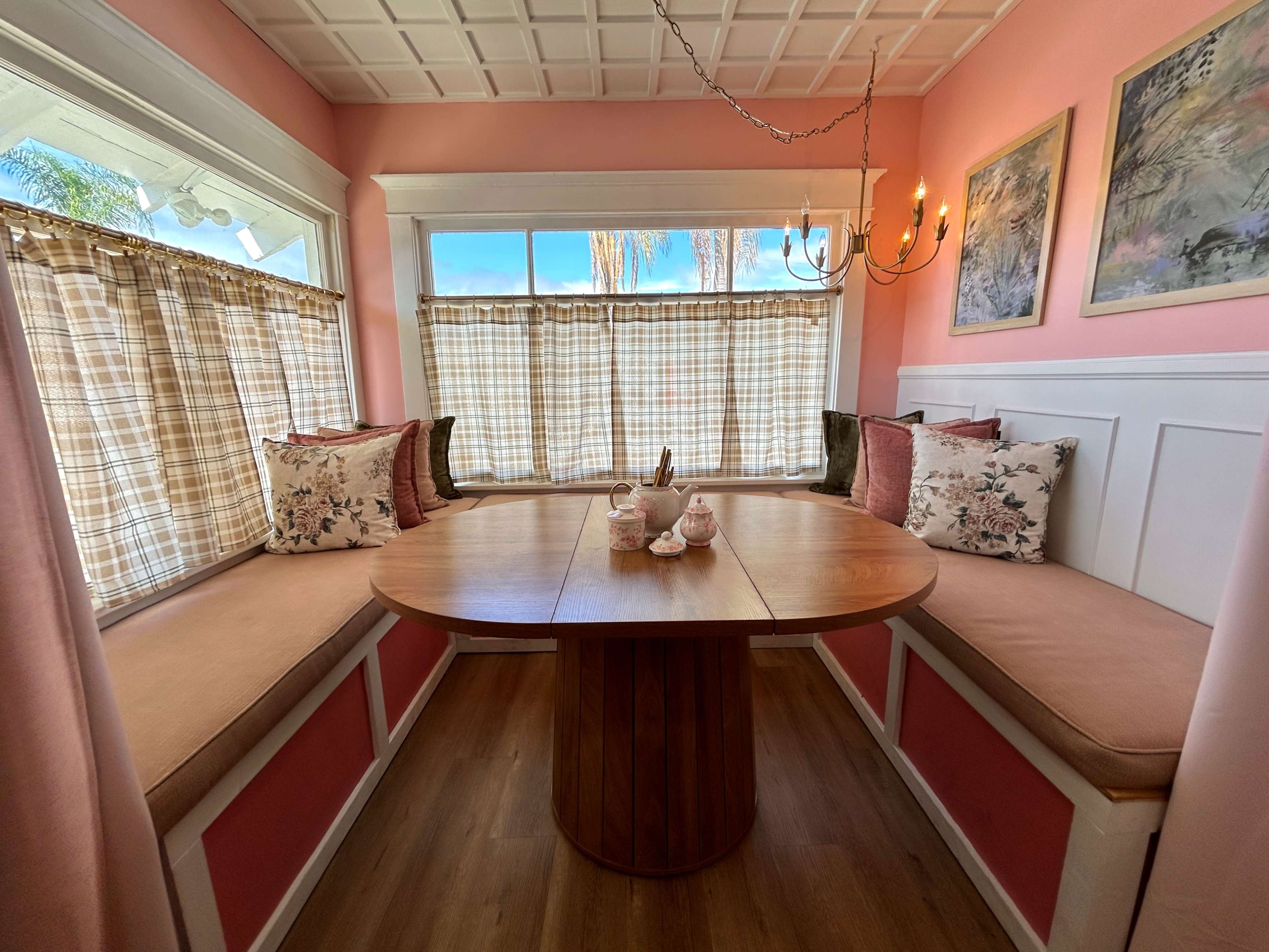 The image shows a cozy dining nook with a round wooden table surrounded by cushioned benches and large windows decorated with checkered curtains.