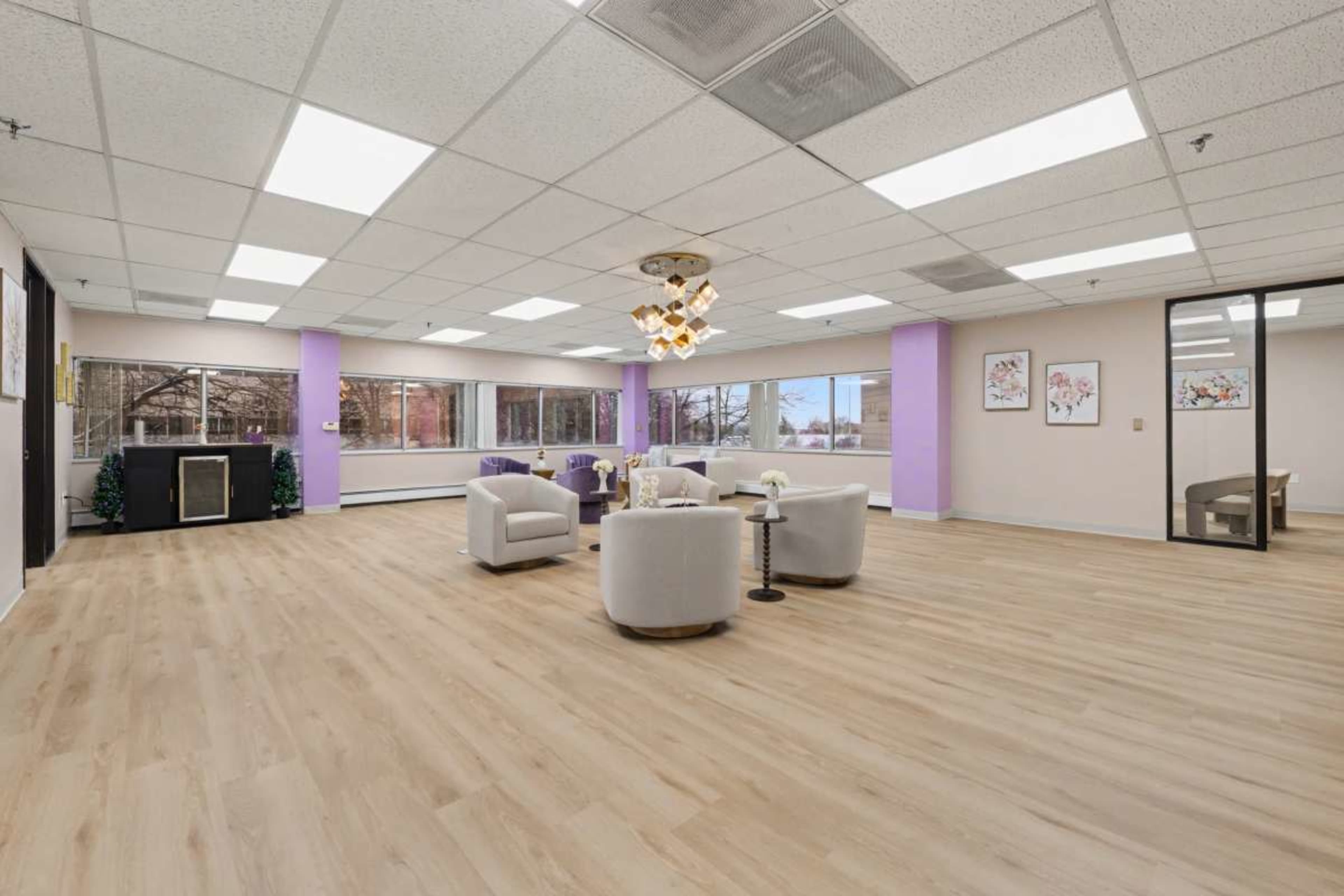 The image shows a spacious, well-lit room with light wood flooring, circular seating arrangements, and large windows.