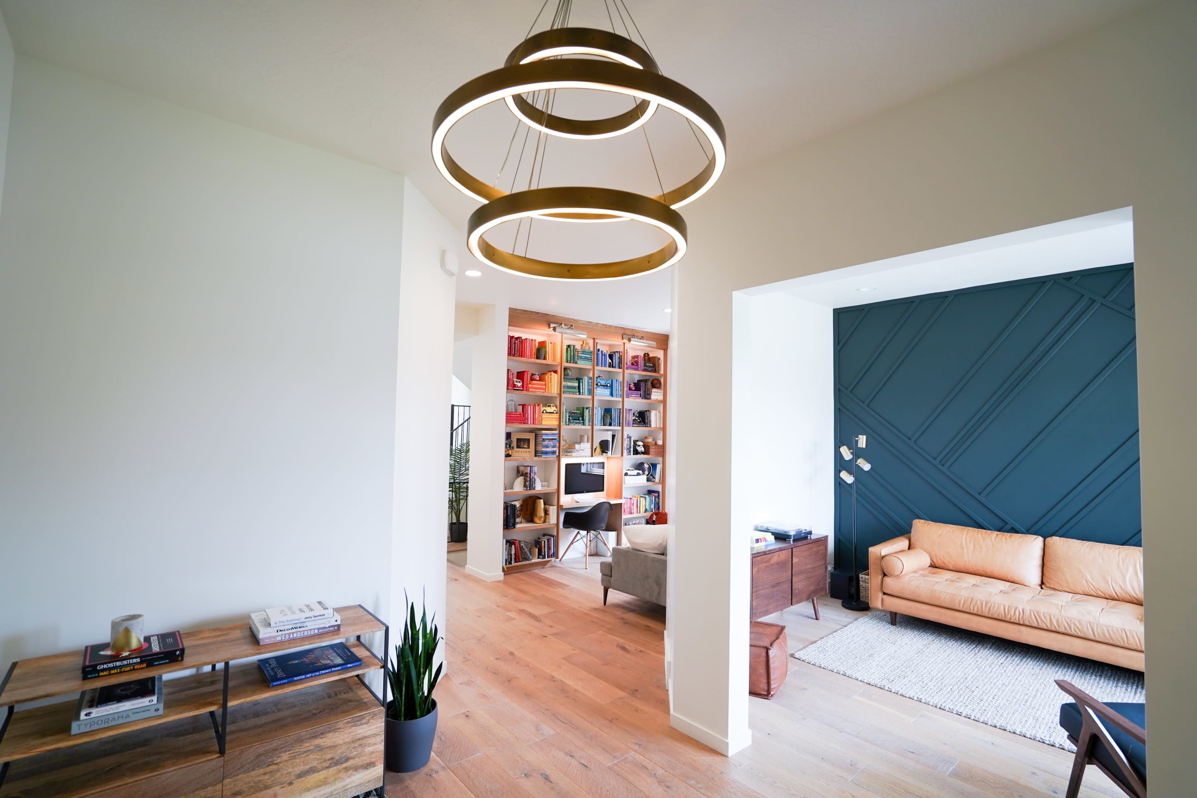 A modern interior space features a large chandelier, wooden floors, a bookshelf filled with colorful books, and a leather sofa in a cozy living area.