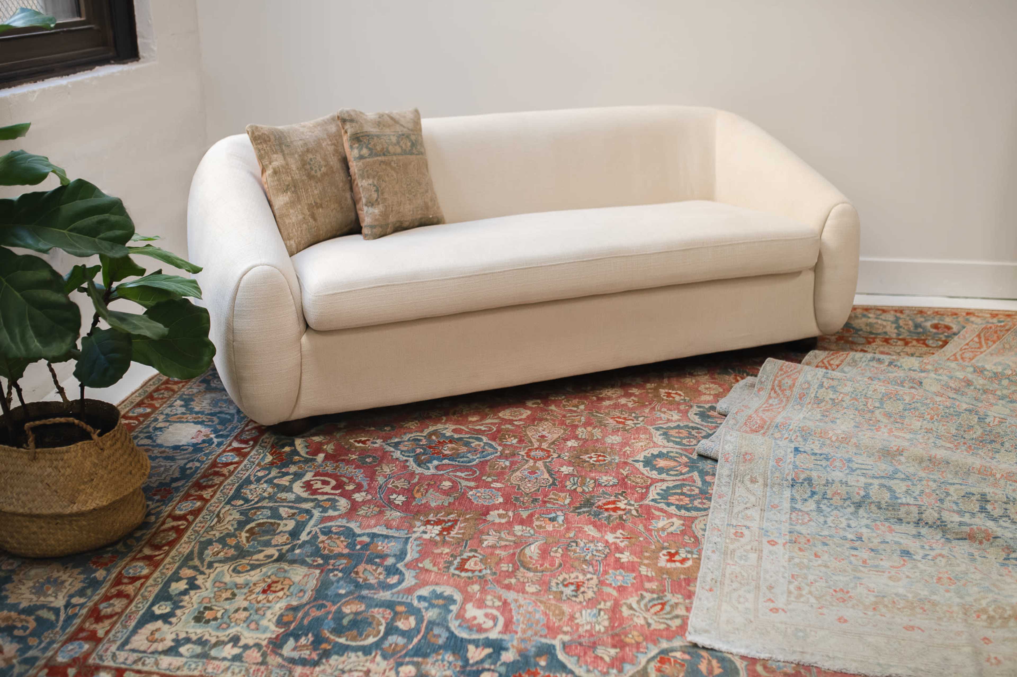 A light-colored, curved sofa with a decorative pillow sits on a patterned rug next to a potted plant in a bright room.
