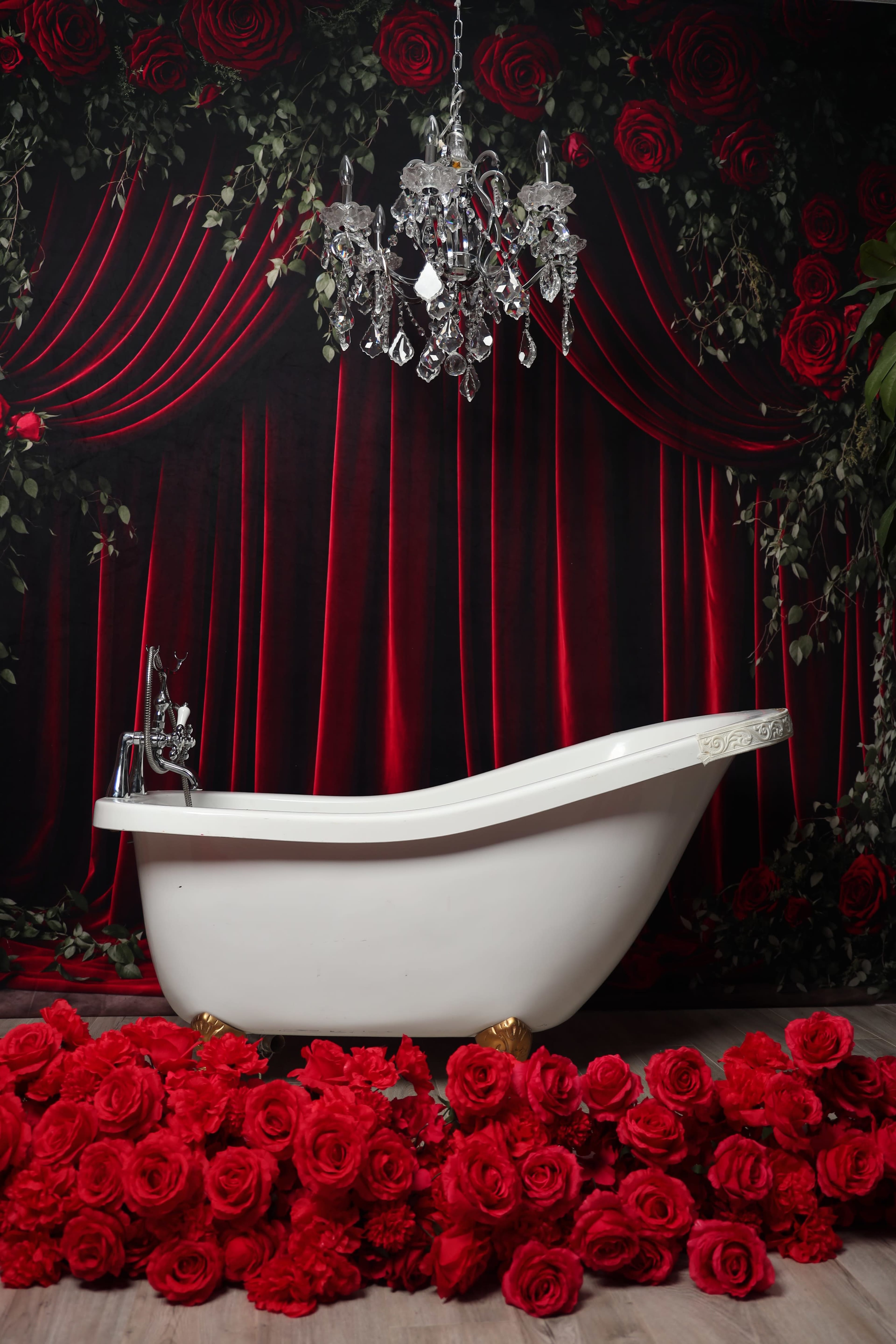 A white freestanding bathtub is positioned in front of a backdrop of red curtains and surrounded by a mound of red roses.