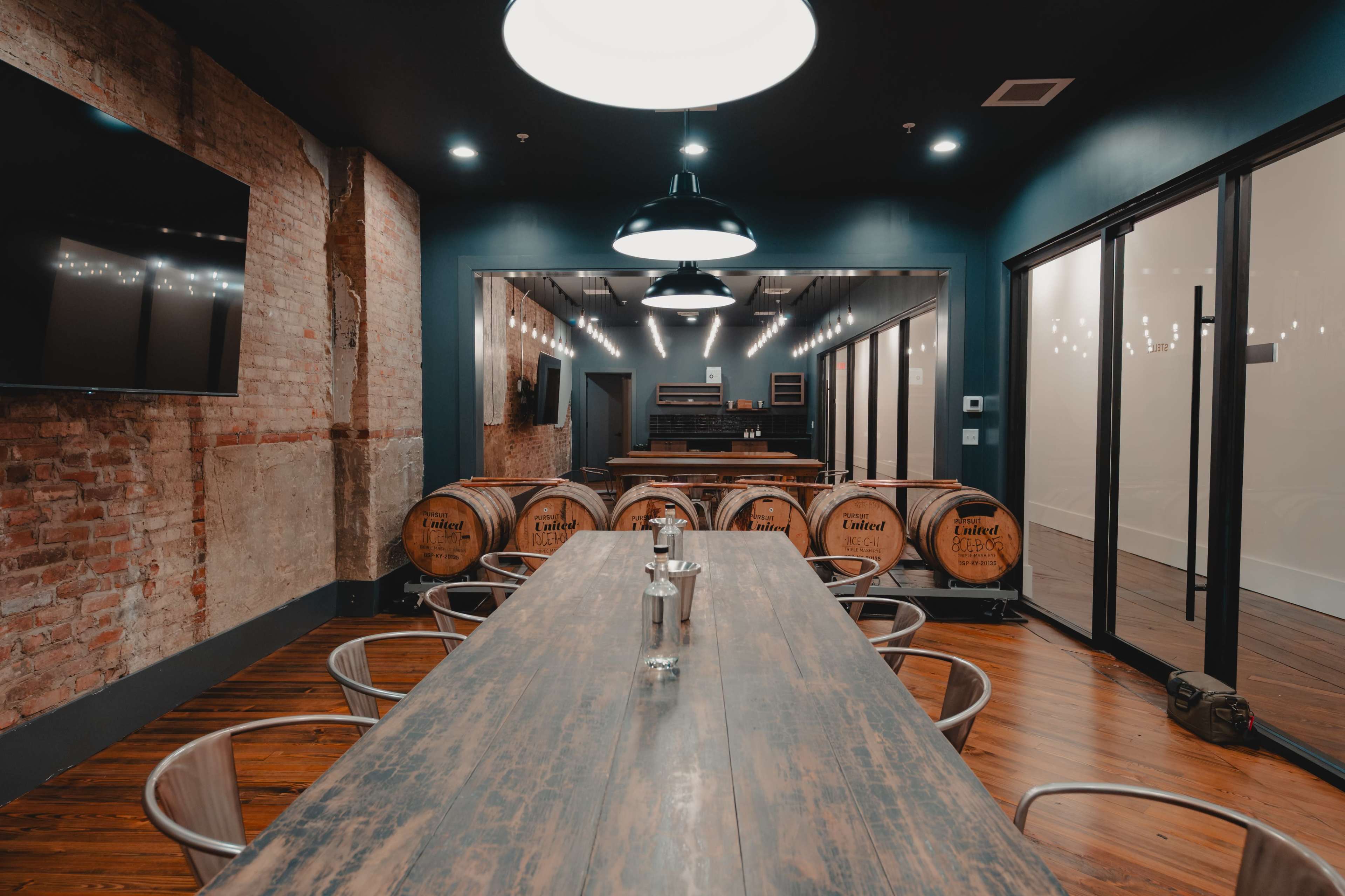 The image shows a modern meeting room with a long wooden table, metal chairs, and wooden barrels in the background, featuring exposed brick walls and large overhead lights.