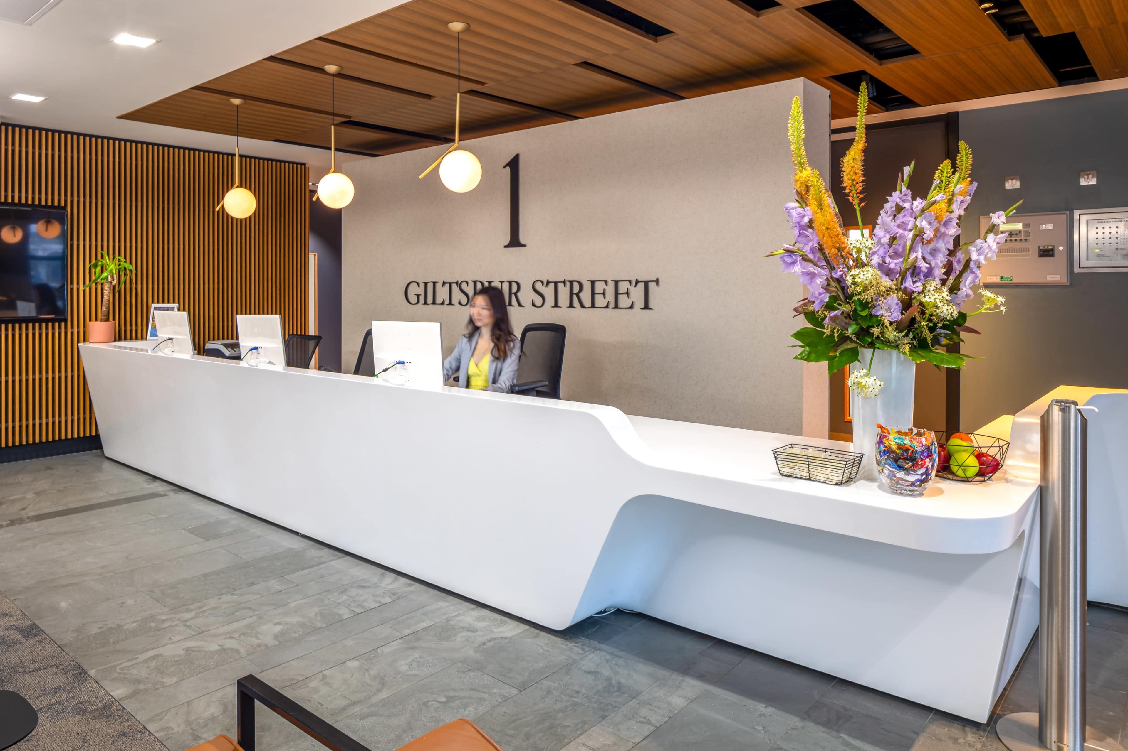 The image shows a modern reception area featuring a sleek white desk, a large floral arrangement, and computer workstations, with the address "1 Giltstar Street" displayed on the wall.