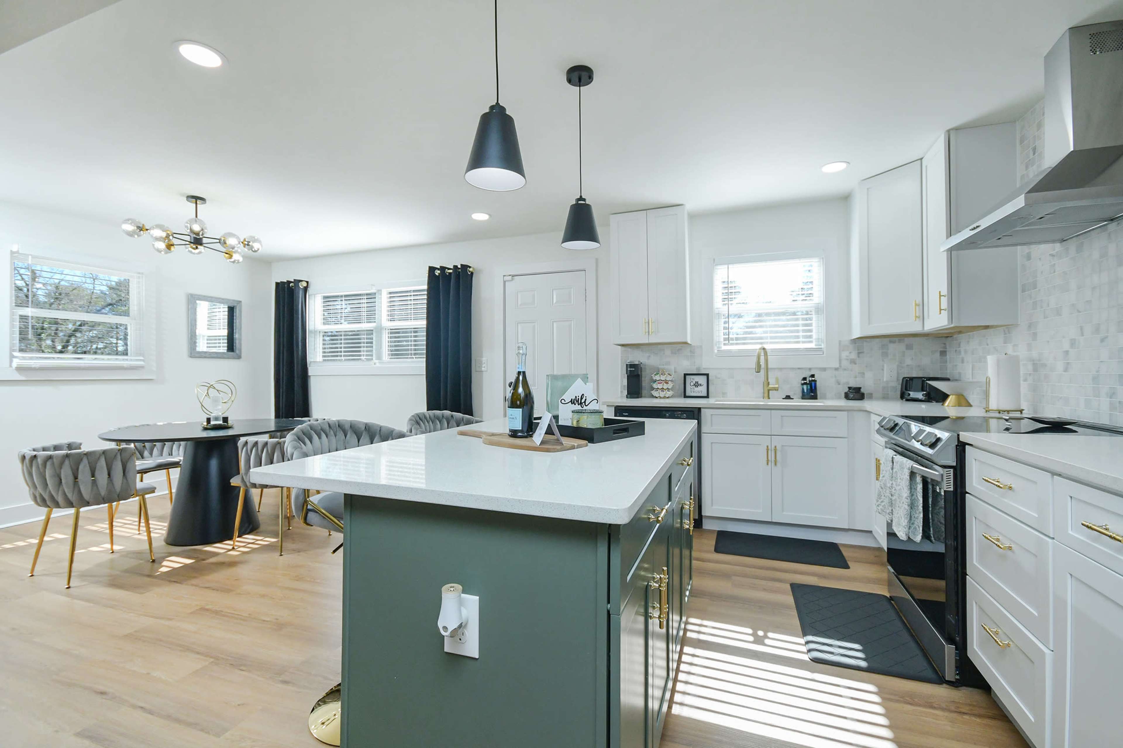 The image shows a modern kitchen with white cabinets, a green island, and a dining area featuring a round table and upholstered chairs.