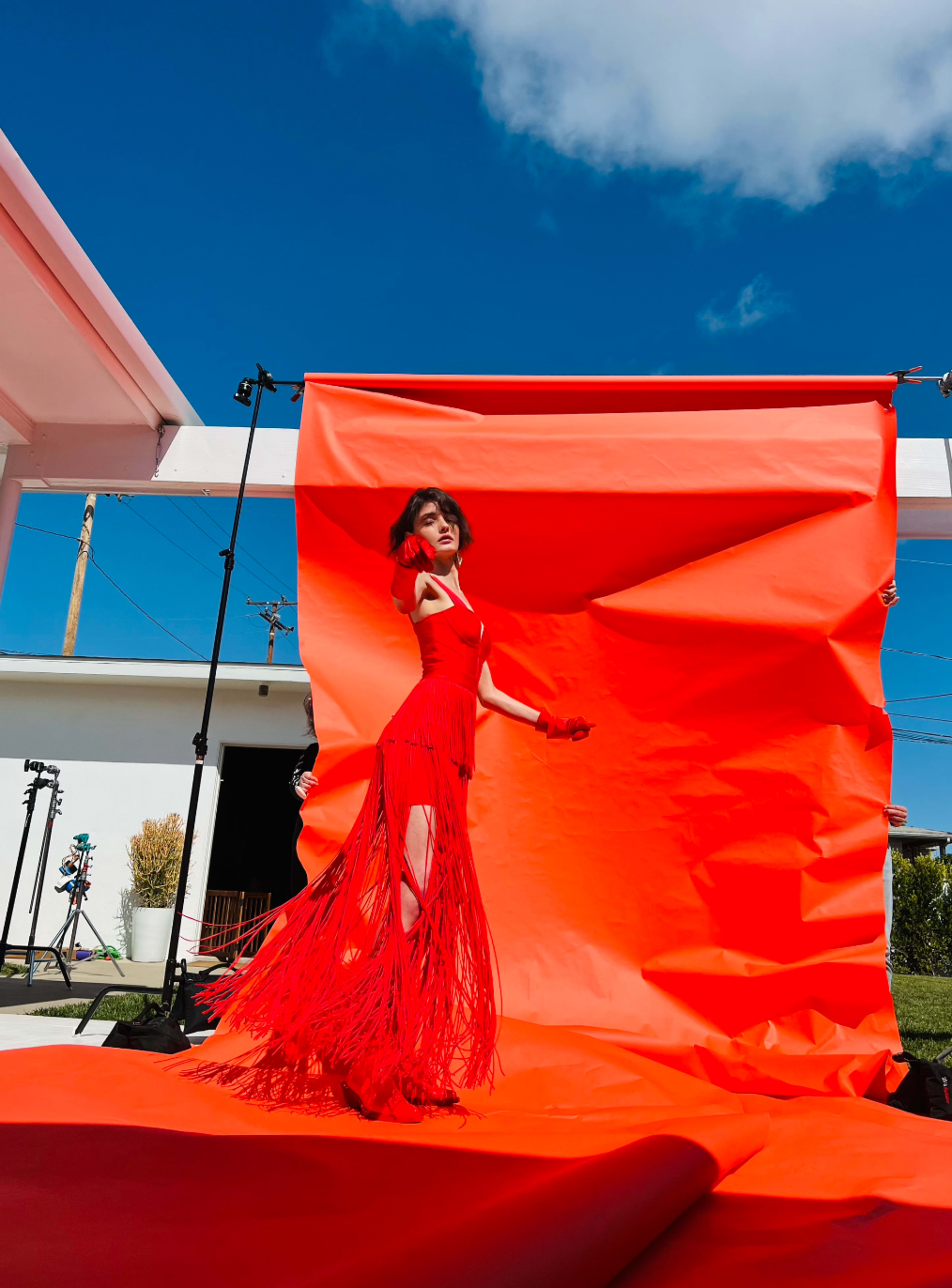 A model poses in a red fringe dress against a bright orange backdrop, with clear blue skies overhead.