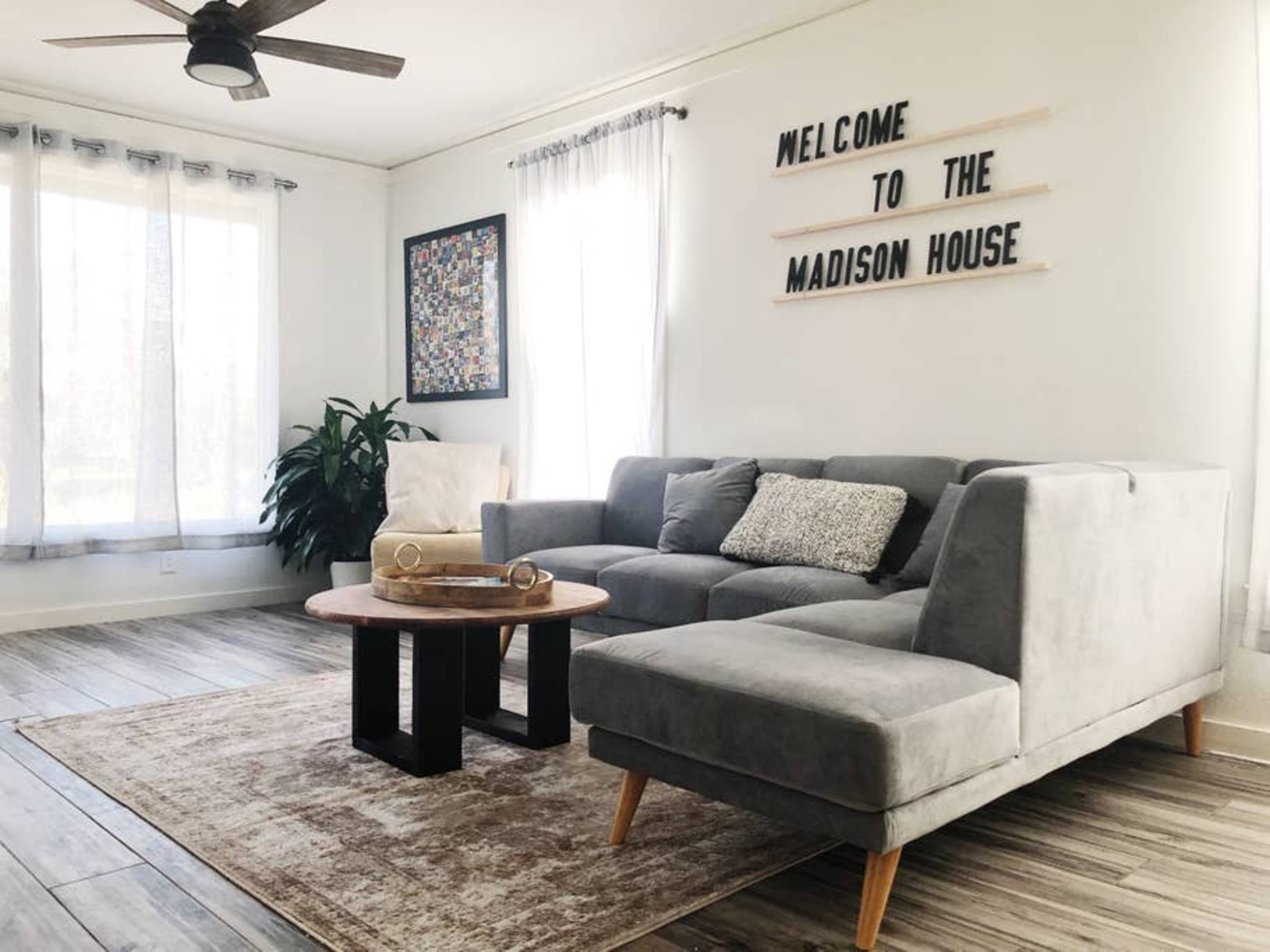 A living room features a gray sectional sofa, a round wooden coffee table, large windows with sheer curtains, and a wall sign that reads "WELCOME TO THE MADISON HOUSE."