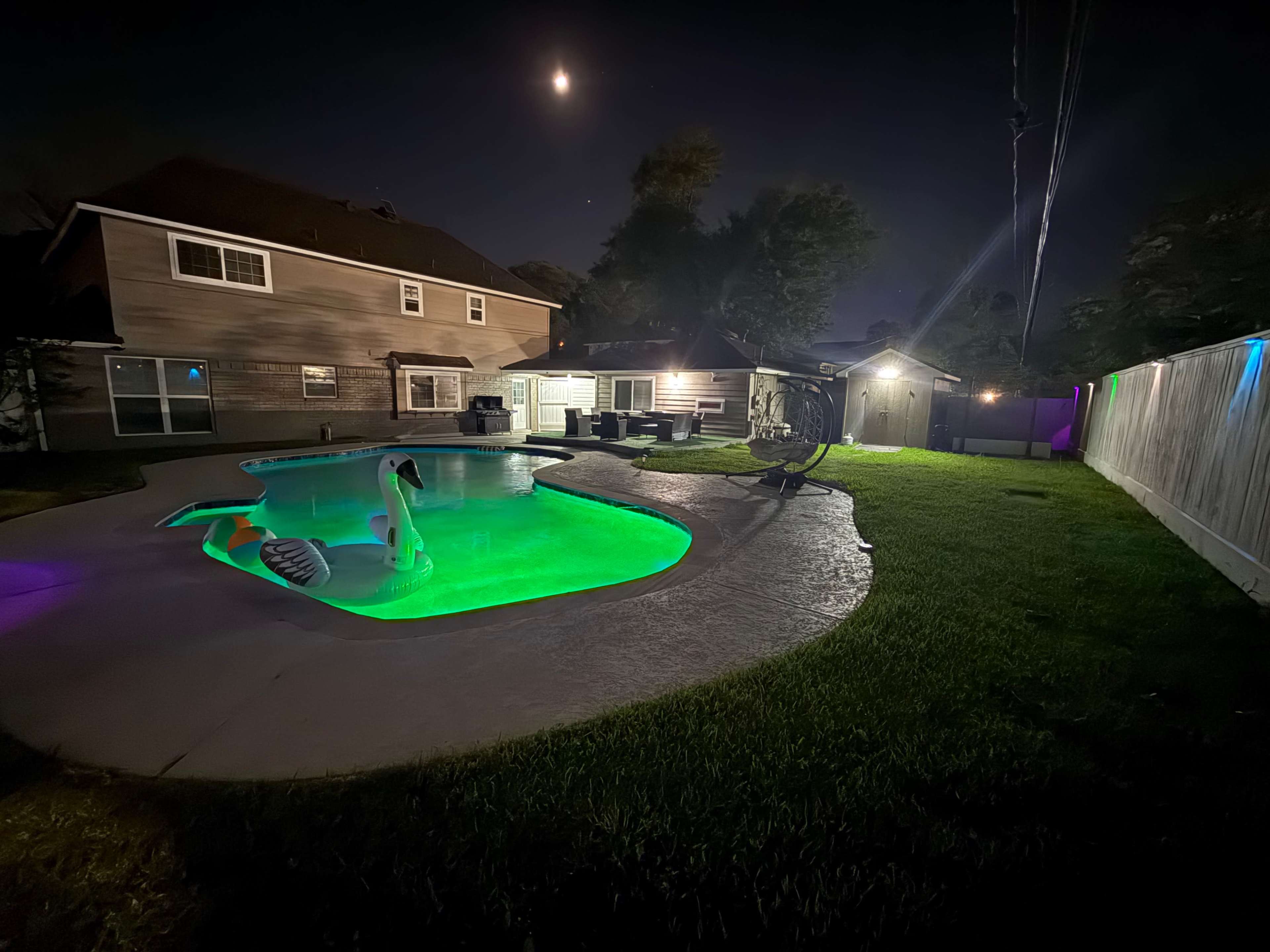 A well-lit backyard features a swimming pool with green illumination, a swan-shaped float, and a house in the background under a clear night sky.