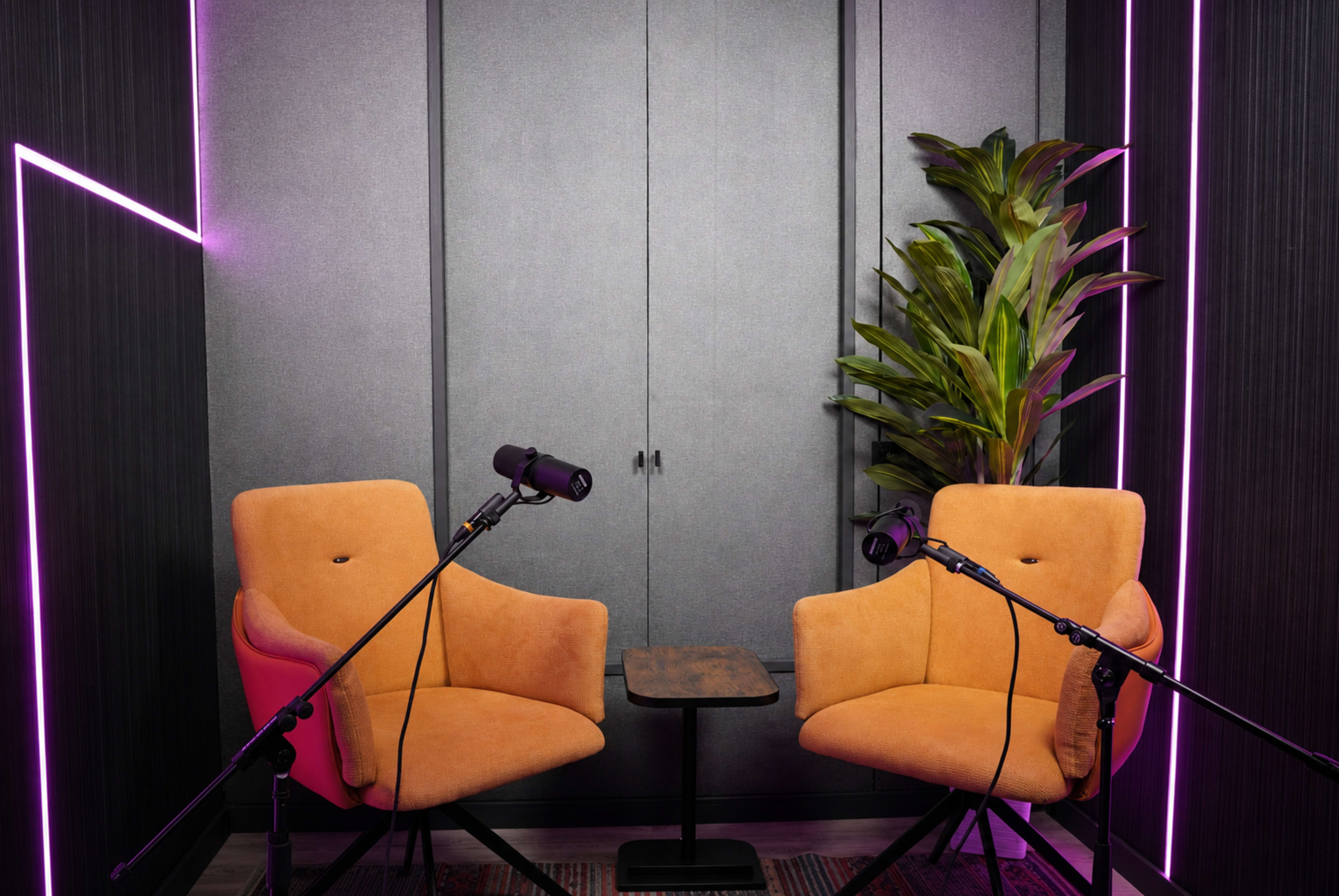 The image shows a podcasting setup featuring two orange chairs, a small table between them, and microphones mounted on stands against a backdrop of gray panels and pink neon lighting.