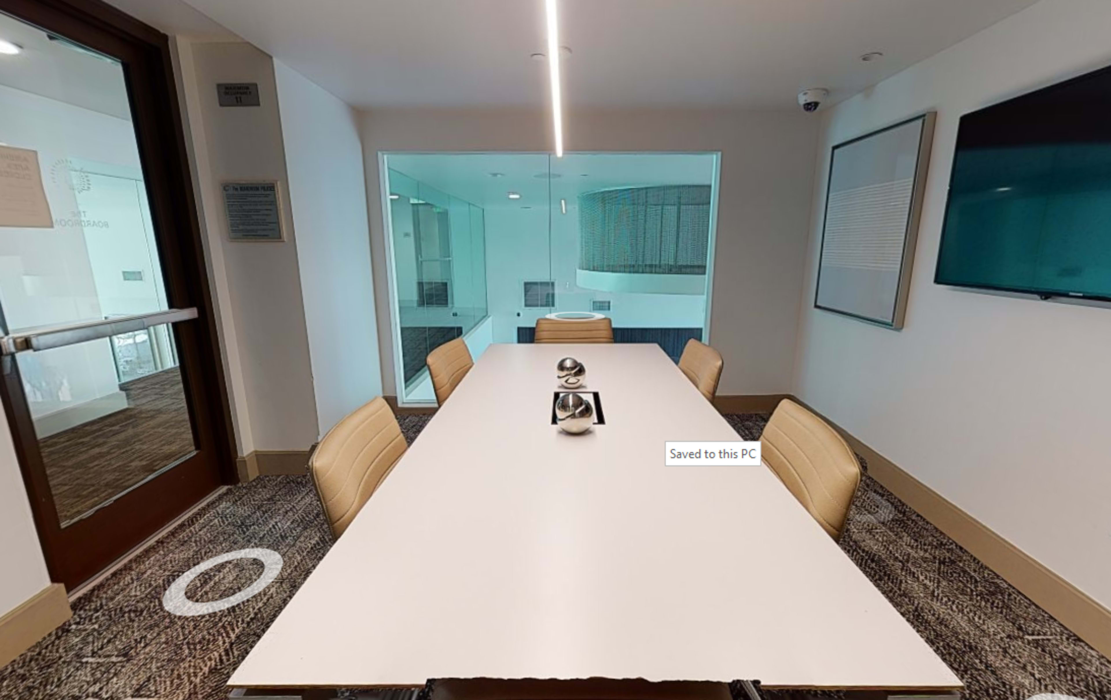 A conference room features a long white table surrounded by beige chairs, with a glass wall showing a view of another room.