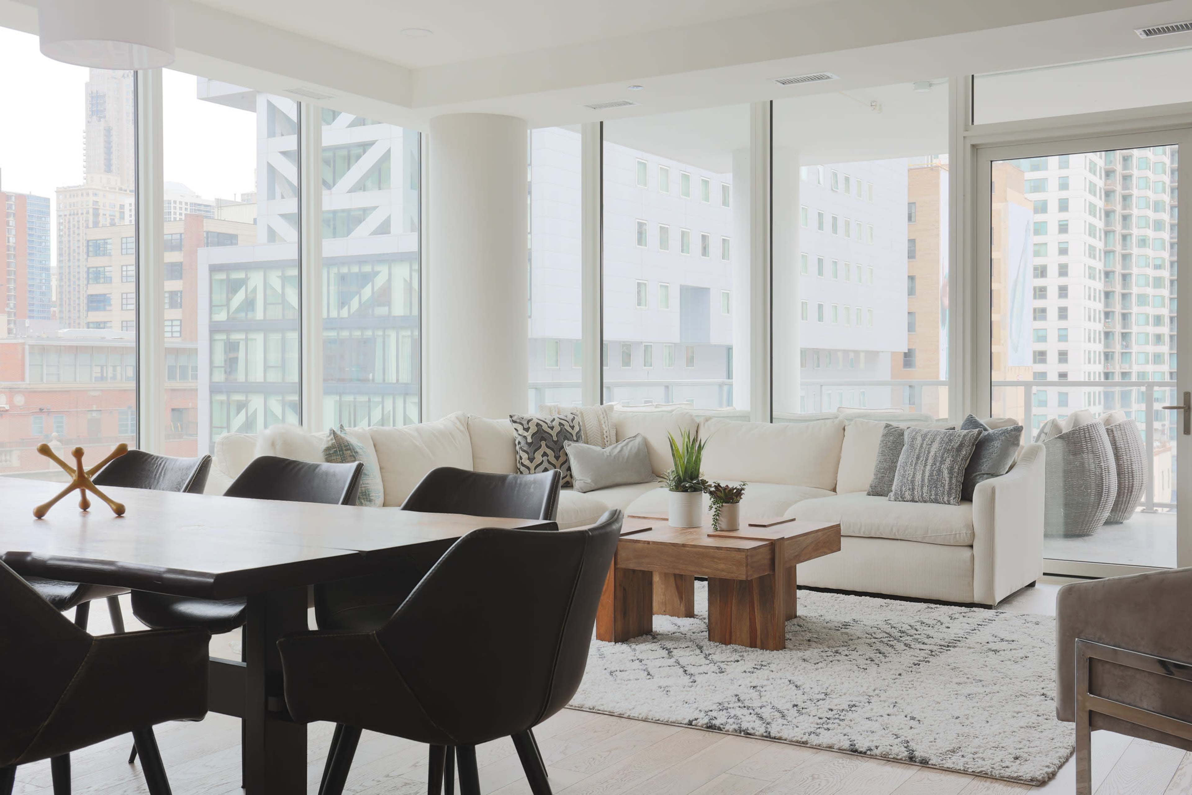 A modern living room features a large sectional sofa, a wooden coffee table, and a dining area with black chairs, surrounded by floor-to-ceiling windows.