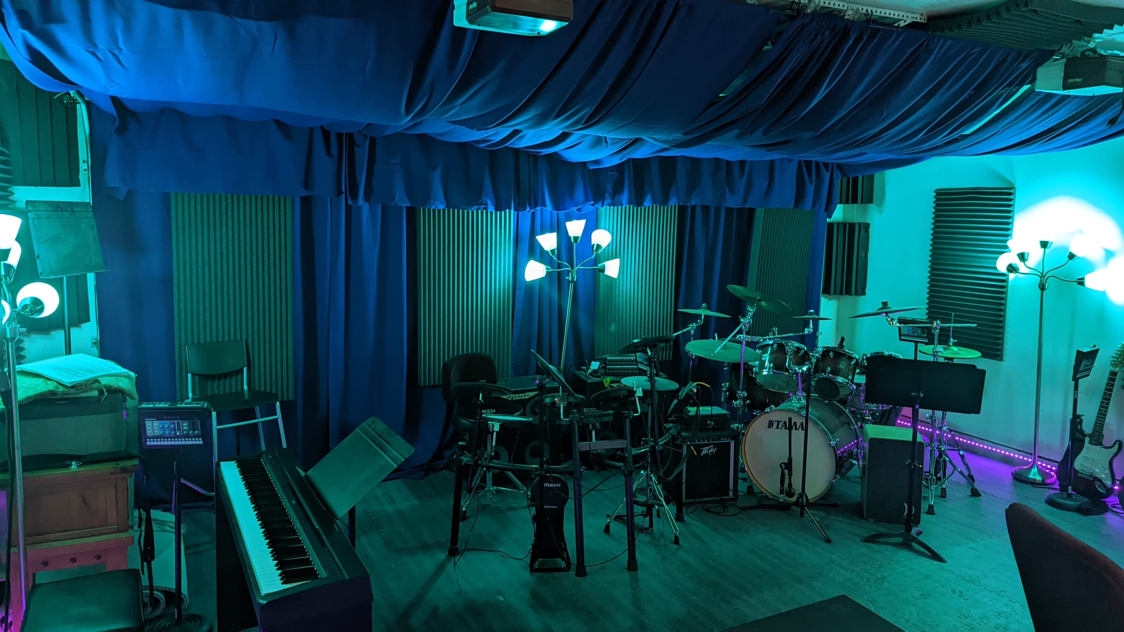 The image shows a music studio featuring a drum set, electric instruments, a piano, and various lighting elements against blue fabric drapes.