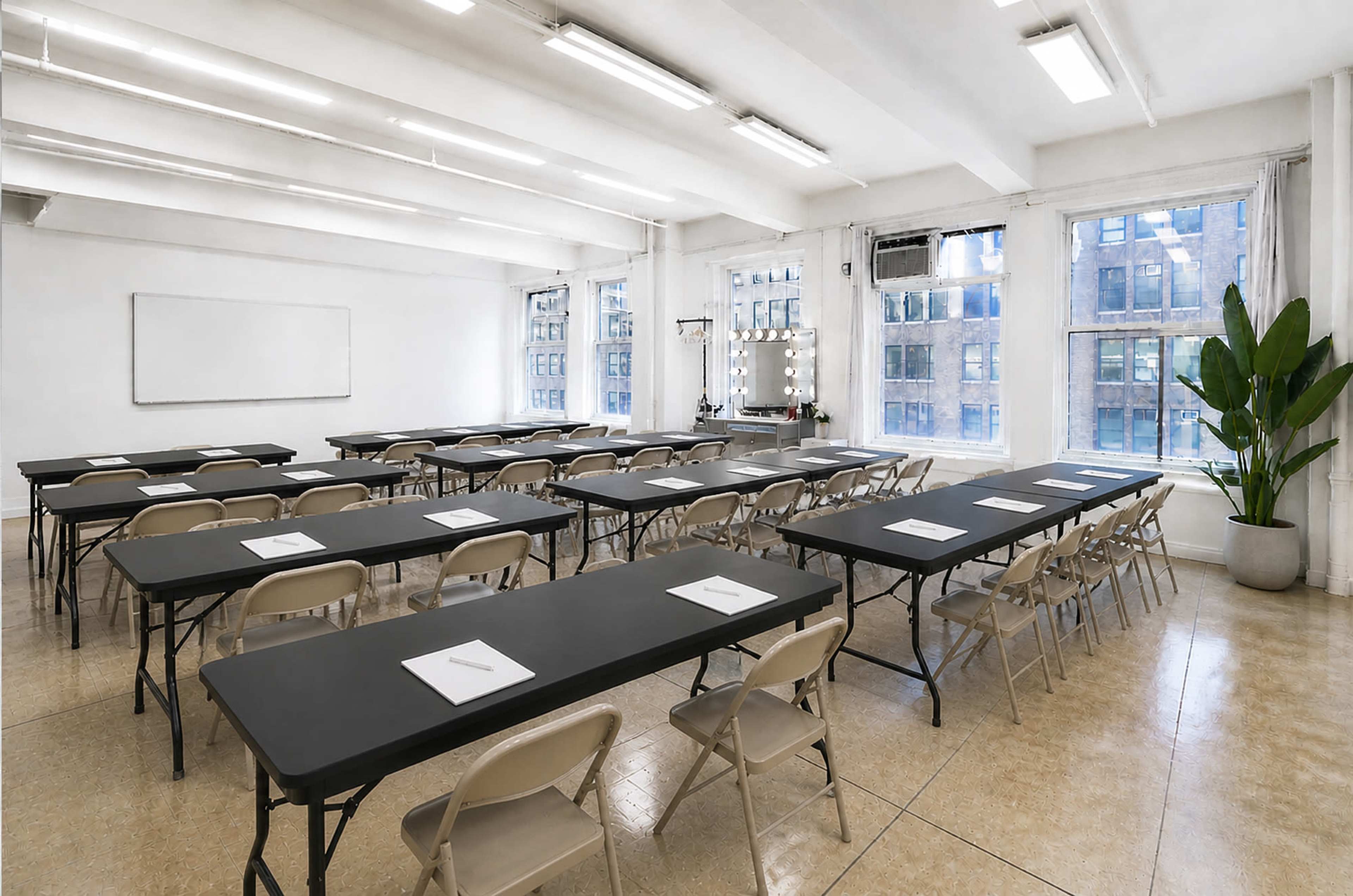 The image shows a spacious classroom set up with multiple rows of black tables and beige chairs, illuminated by large windows.
