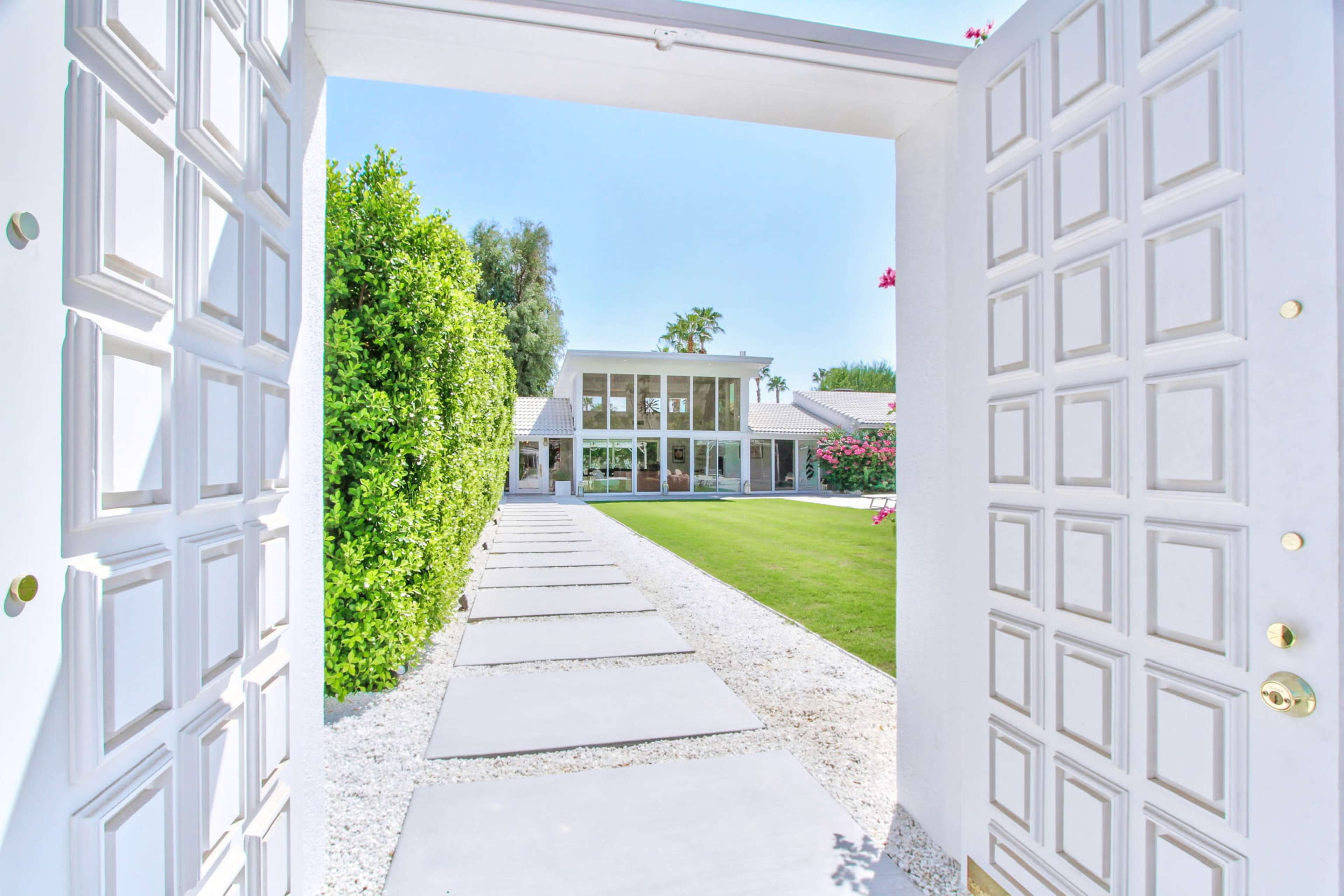 A modern home is visible through an open white front door, with a pathway lined by stone pavers leading to a green lawn and landscaped shrubs.
