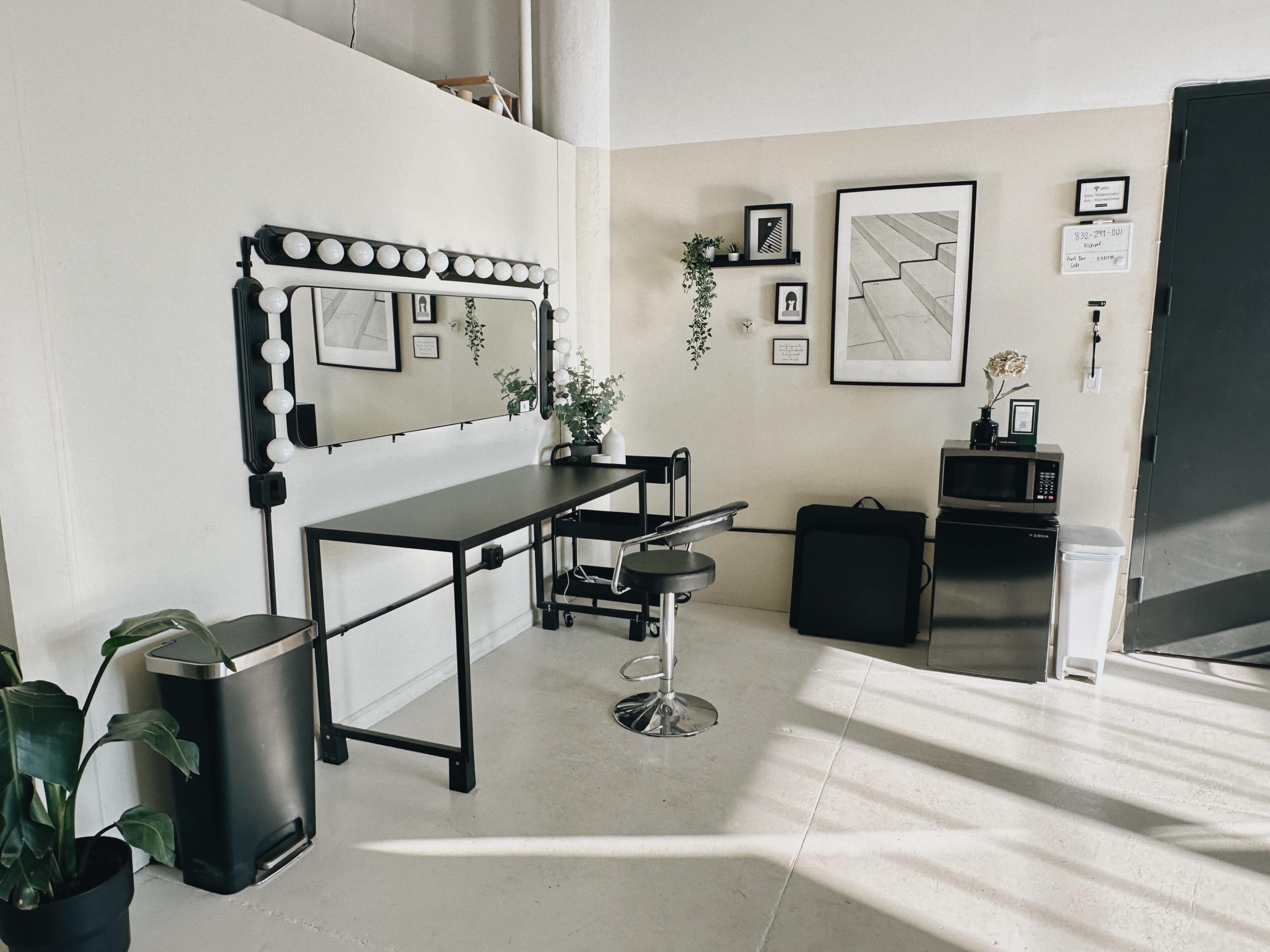 The image shows a minimalist dressing area with a lighted mirror, a black table and chair, framed artwork on the walls, and a microwave and mini fridge in the corner.