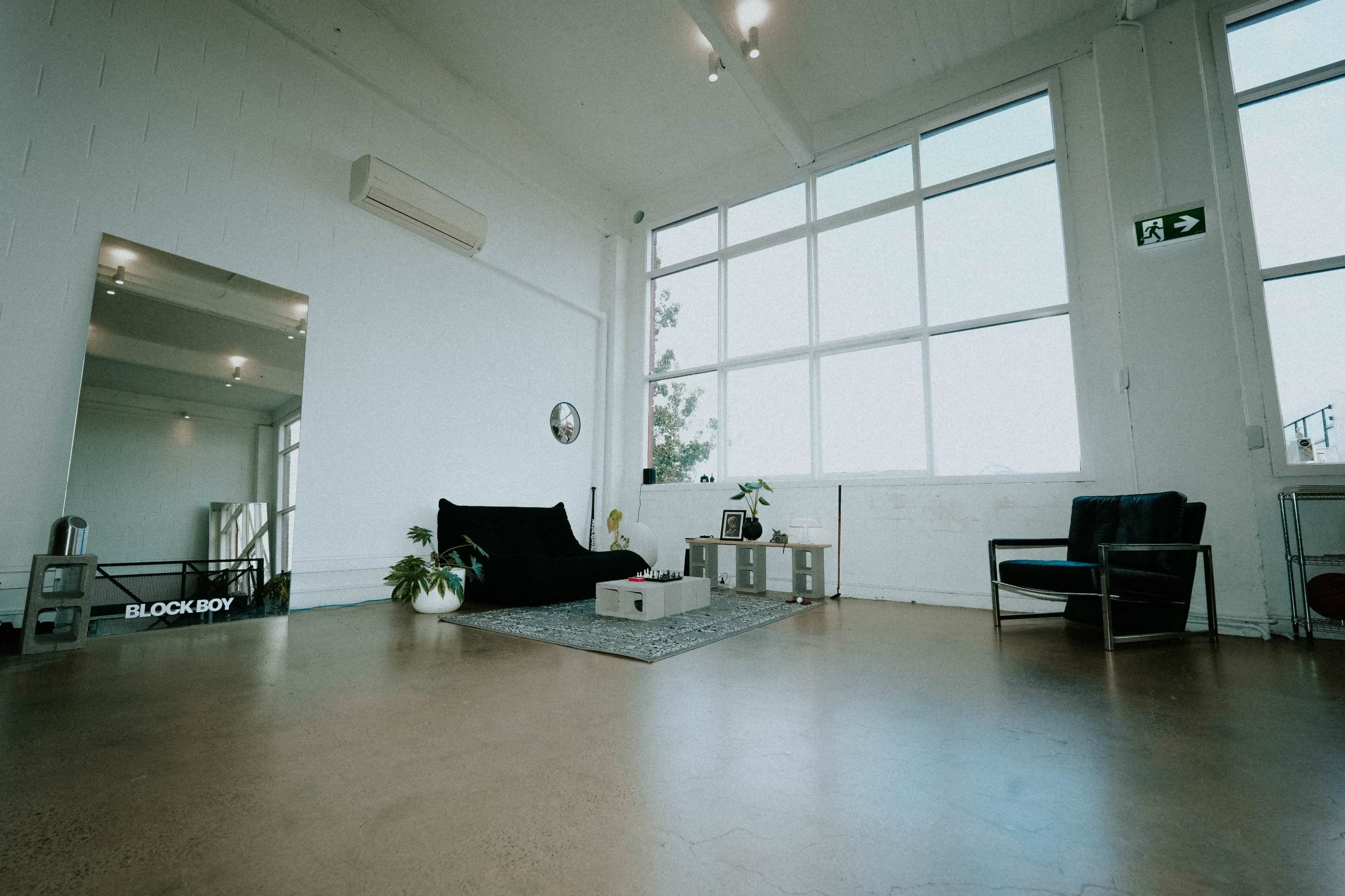 The spacious interior features large windows, a black couch, a black chair, a coffee table, and a decorative rug on a polished concrete floor.