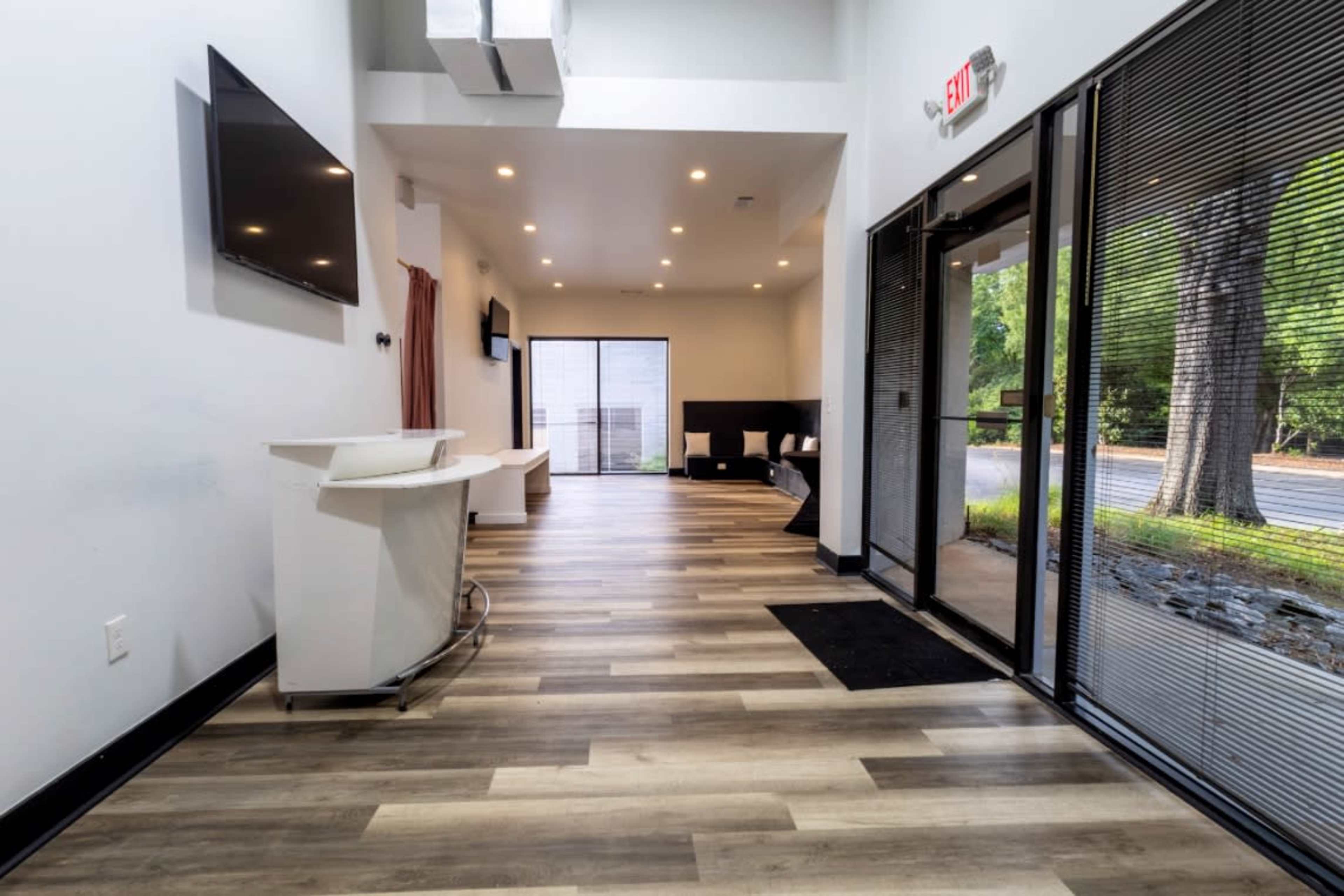 The image shows a modern, open reception area with wooden flooring, large windows, and minimalistic furnishings.