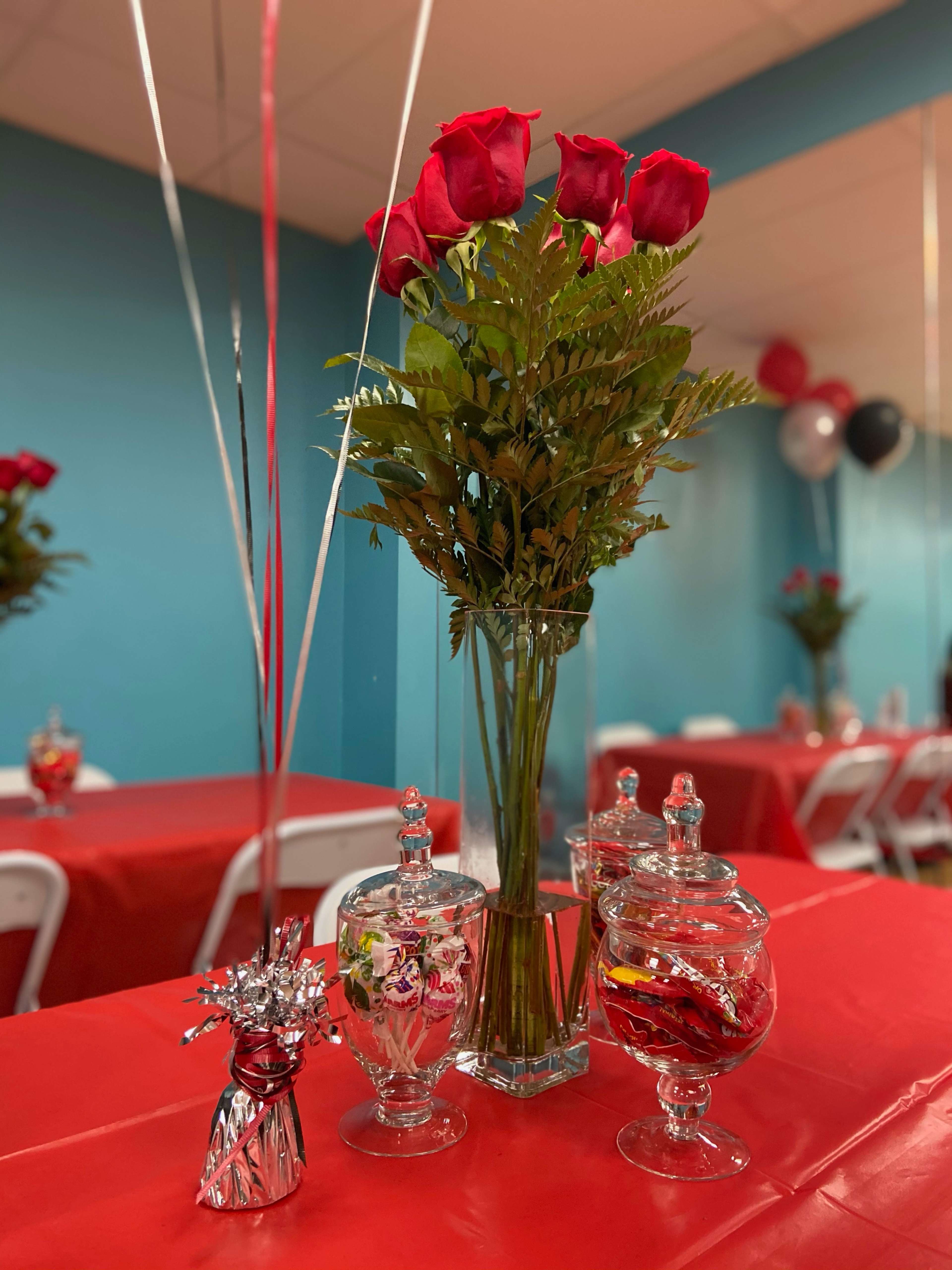 A bouquet of red roses and greenery sits in a clear vase on a table with a red tablecloth, accompanied by decorative glass jars and balloons in a festive setting.