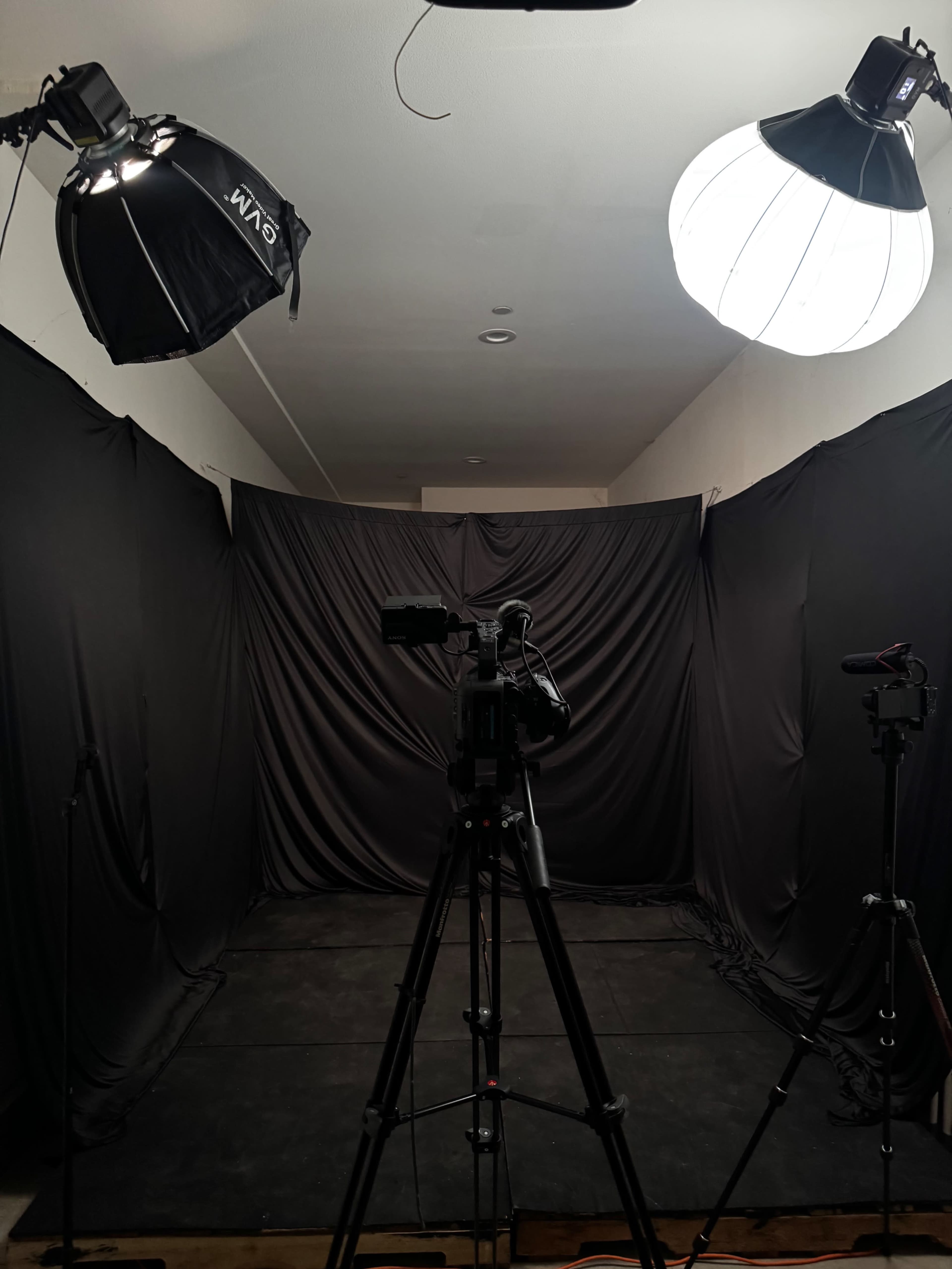 The image shows a photography studio setup featuring two light sources and a camera on a tripod facing a black backdrop.