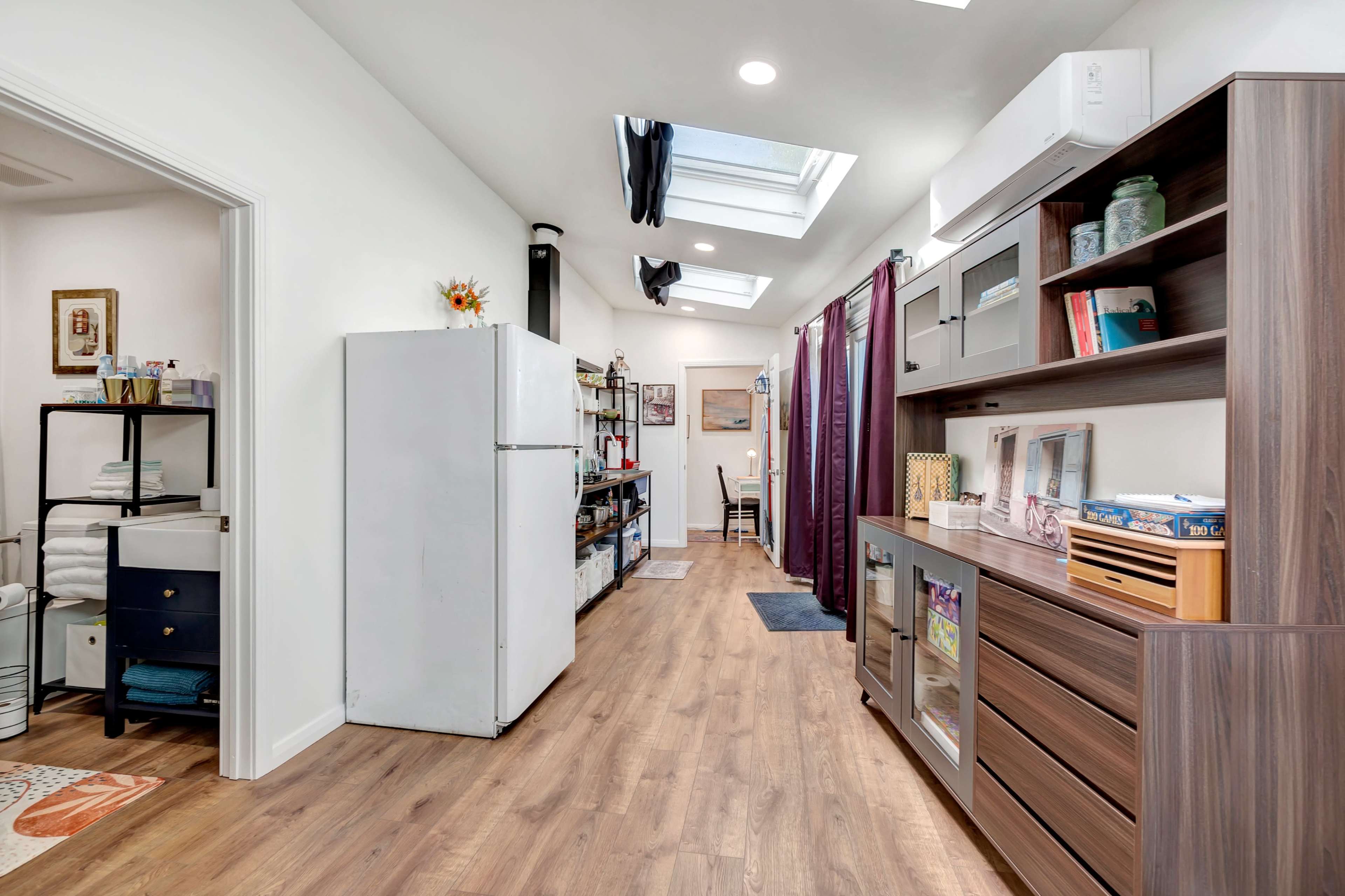 The image shows a modern interior hallway featuring a refrigerator, bookshelves, and natural light from skylights.