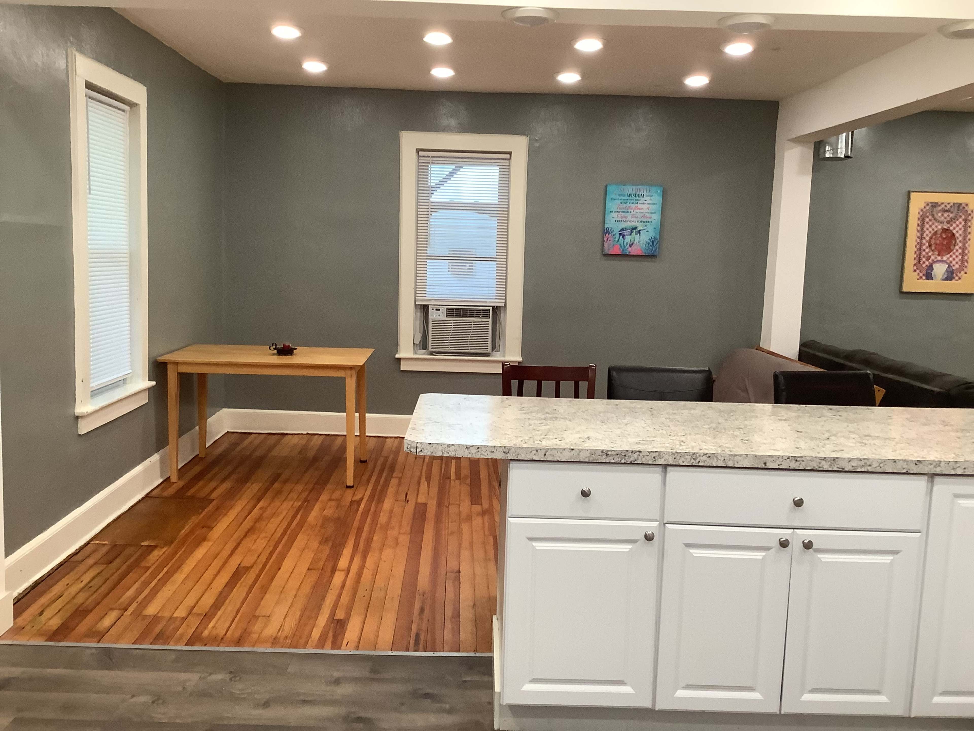 A kitchen area with a white countertop and cabinets, adjacent to a small dining table and chairs, set against a wall with light-colored paint and wood flooring.