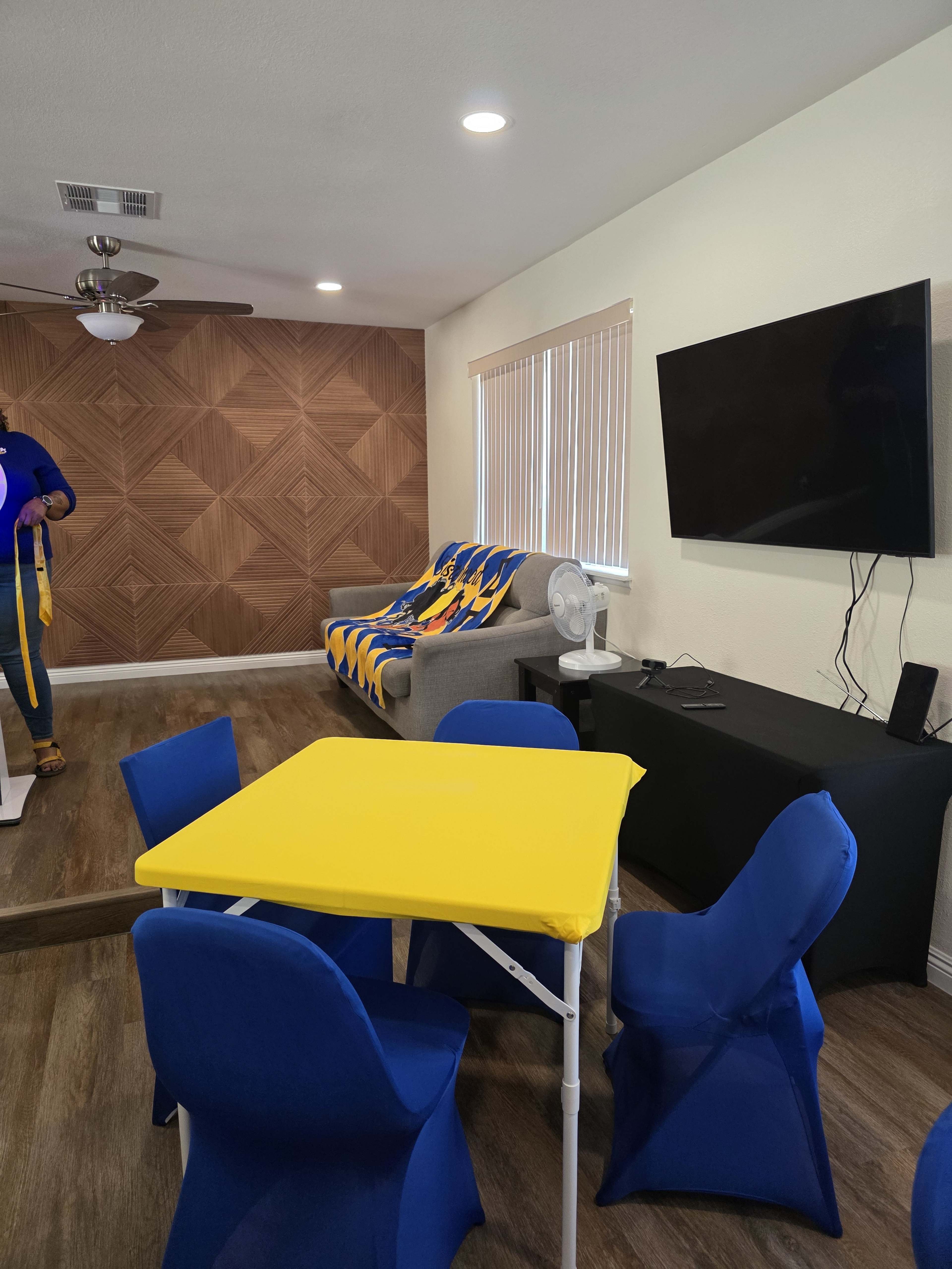 The image shows a living area with a yellow table, blue chairs, a gray sofa covered in a colorful throw, a fan, and a wall-mounted television.