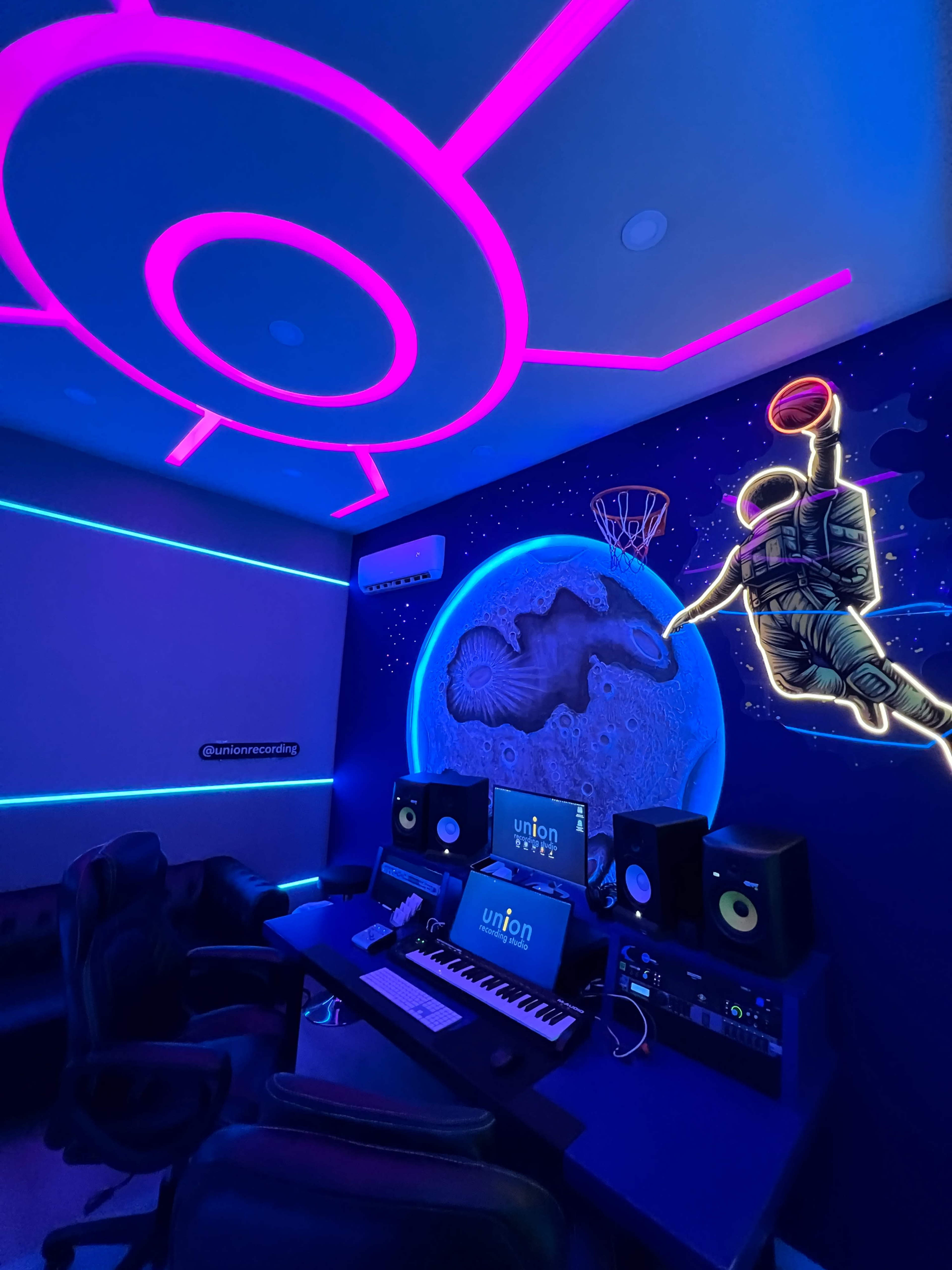 The image shows a modern music studio featuring a desk with audio equipment, neon lighting, and a mural of an astronaut playing basketball against a cosmic background.