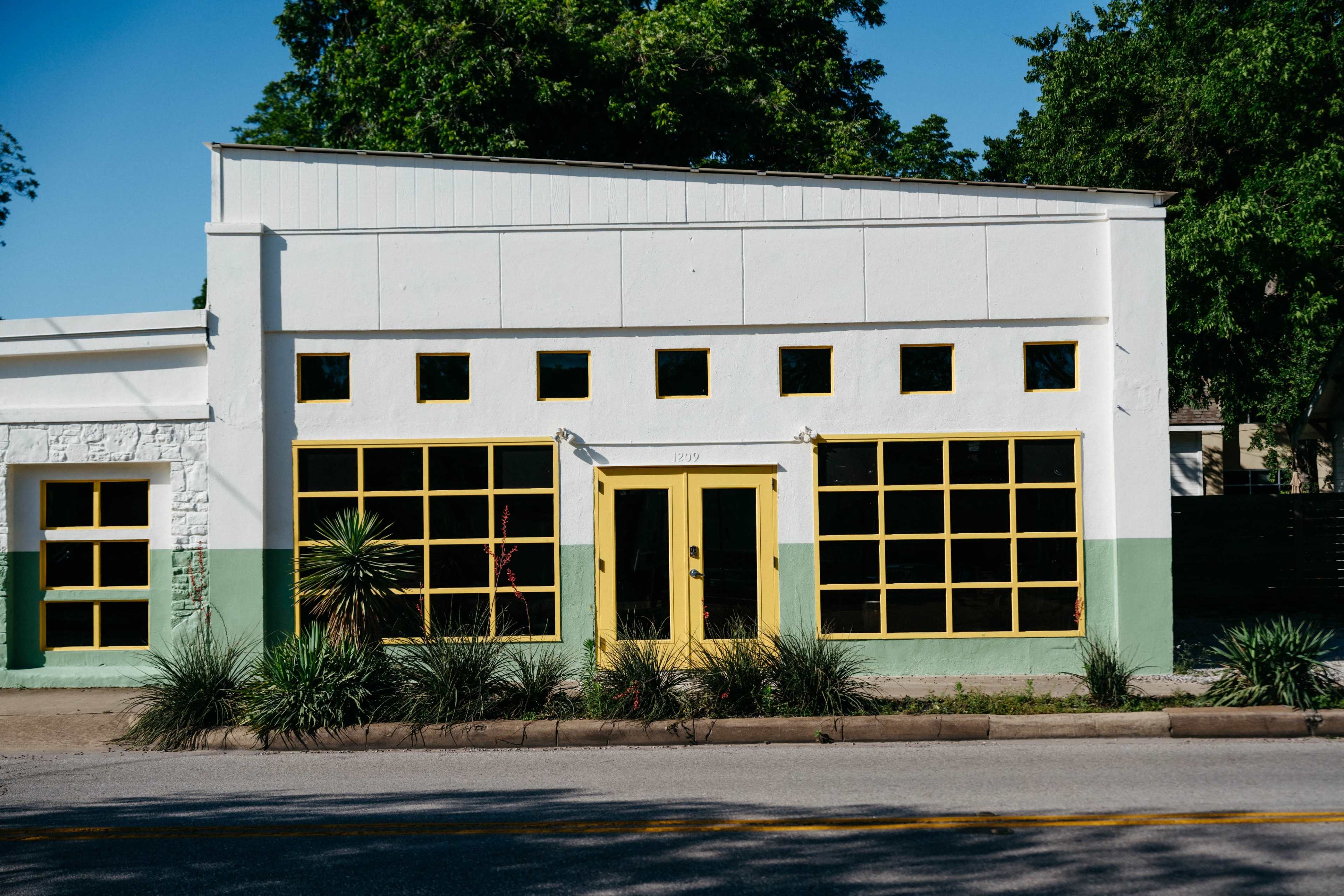The building features a white exterior with yellow trim and large windows on the front facade, set against a backdrop of greenery.