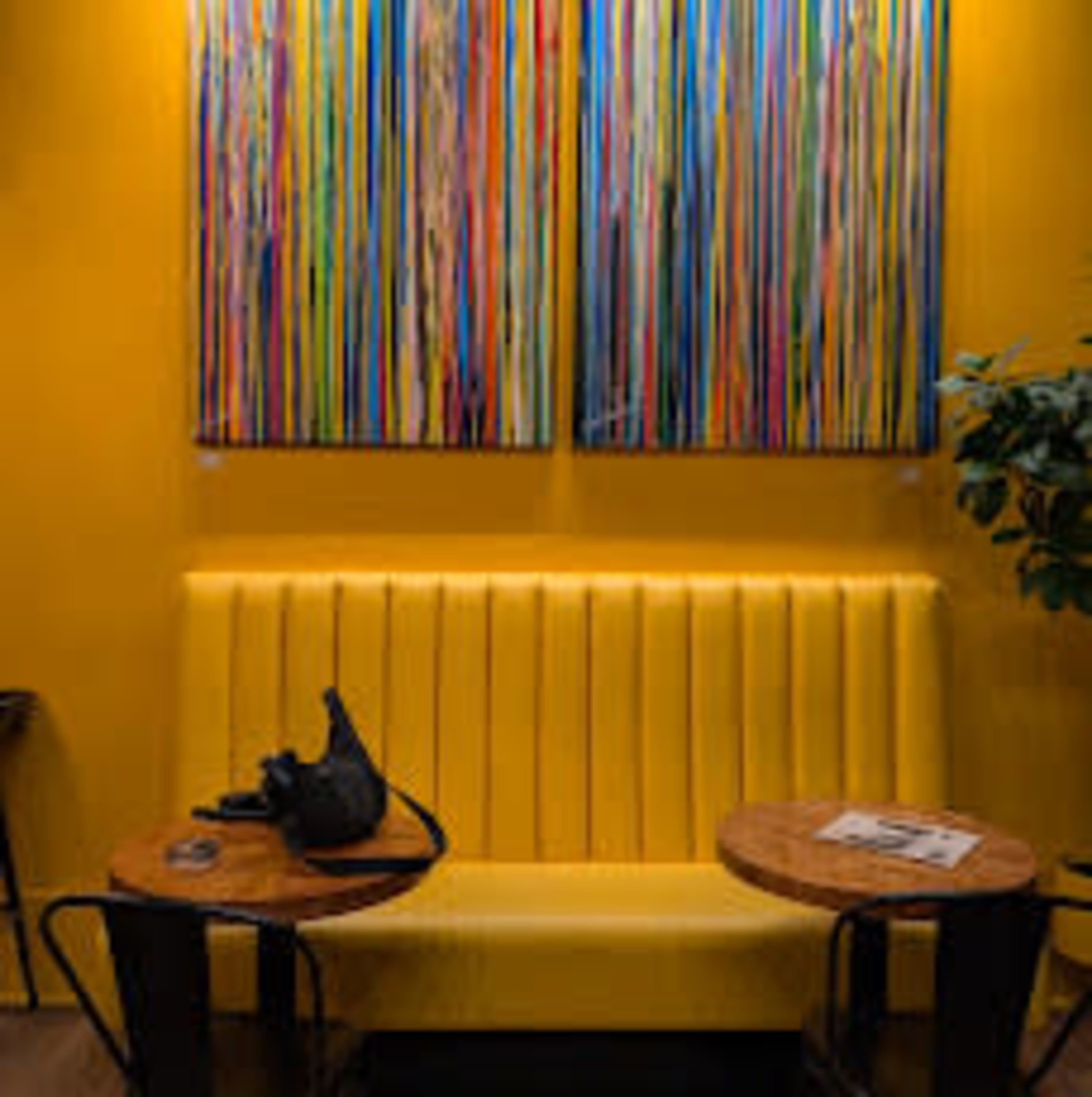 The image shows a bright yellow seating area with a modern tufted bench, two wooden tables, and colorful abstract paintings hanging on the wall.