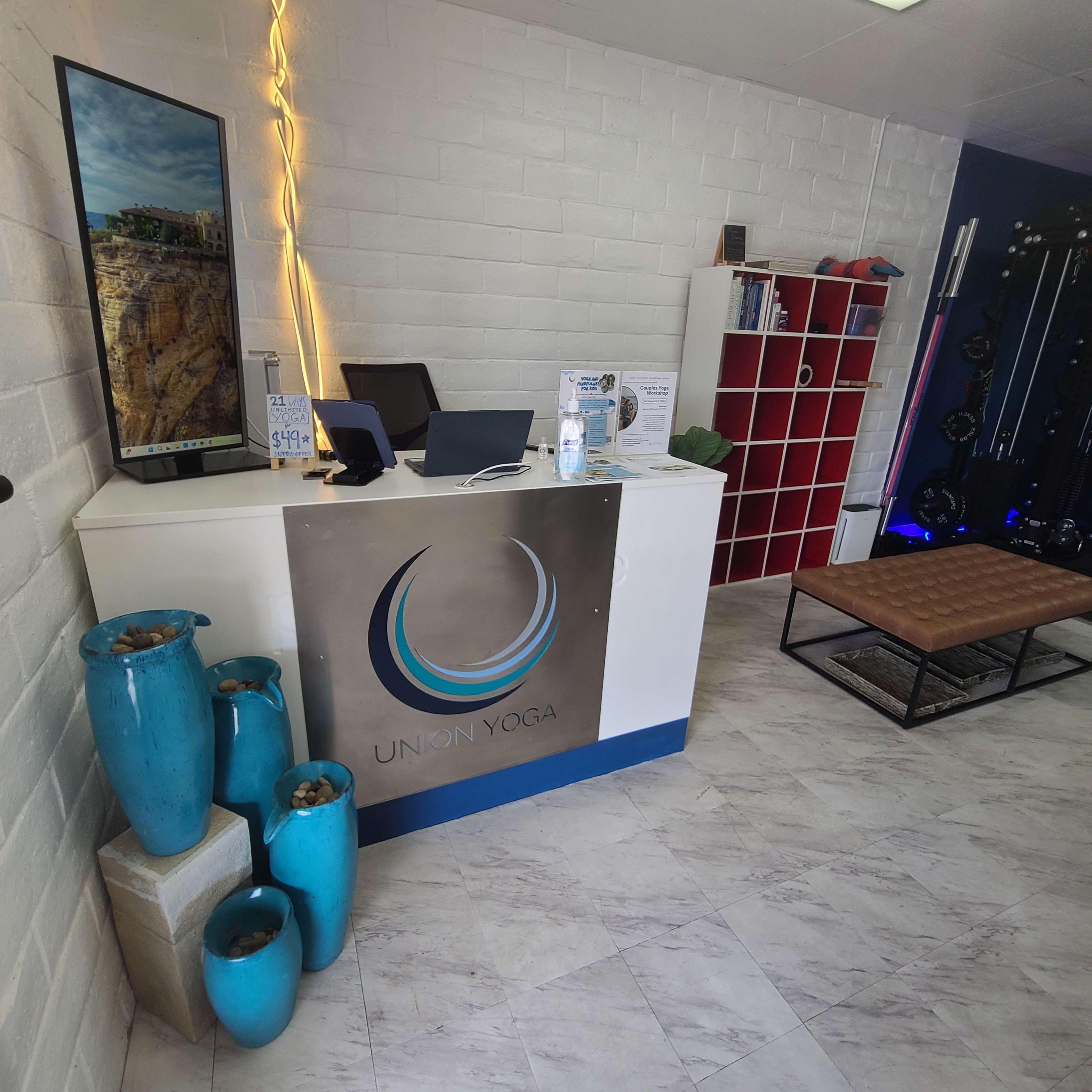 The image shows a reception area of a yoga studio, featuring a desk with a logo, a large framed photo on the wall, blue decorative vases, and exercise equipment in the background.