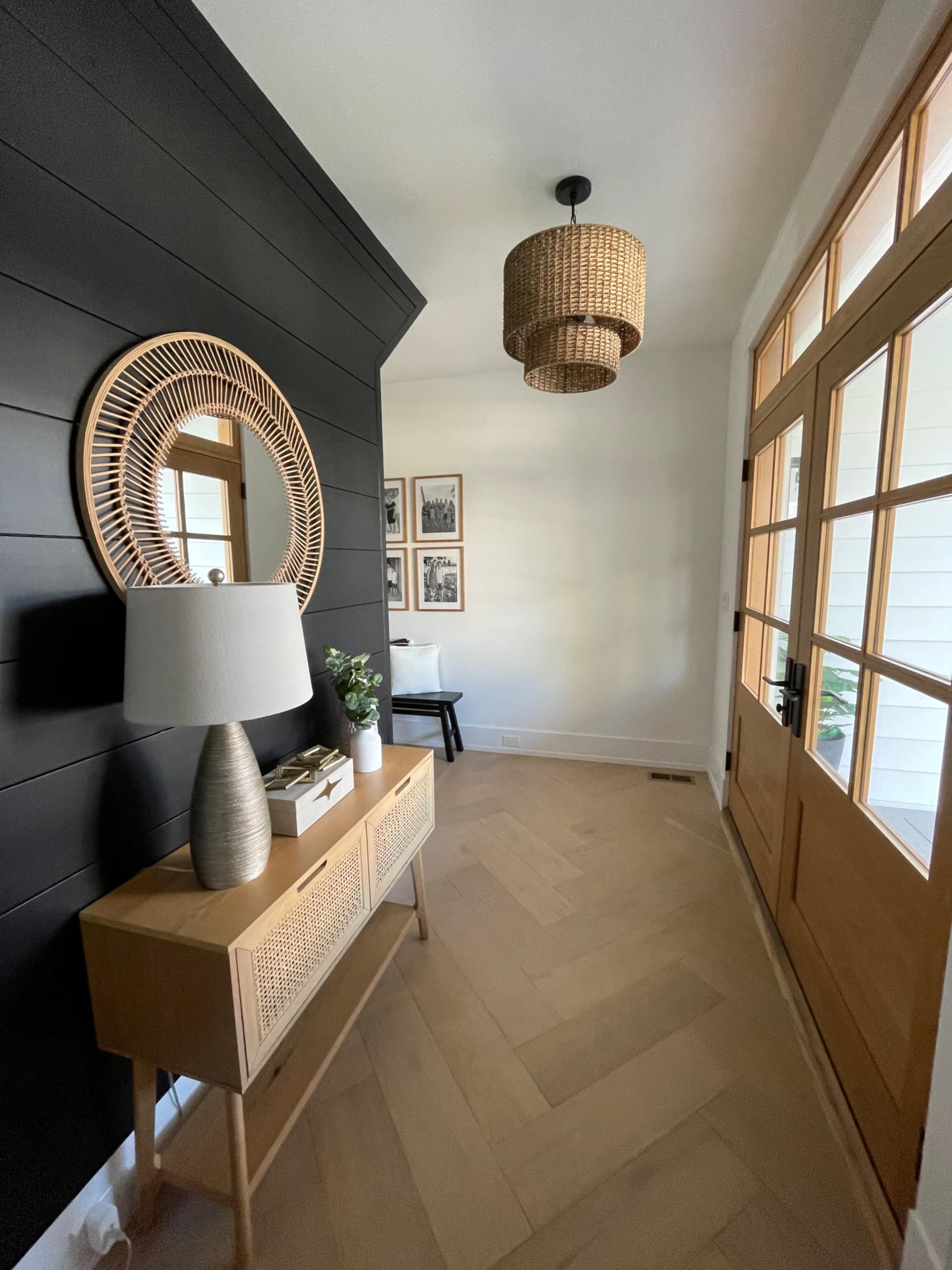 A hallway with a black wall featuring a round mirror, a light fixture hanging from the ceiling, and a console table with decorative items next to a double front door.