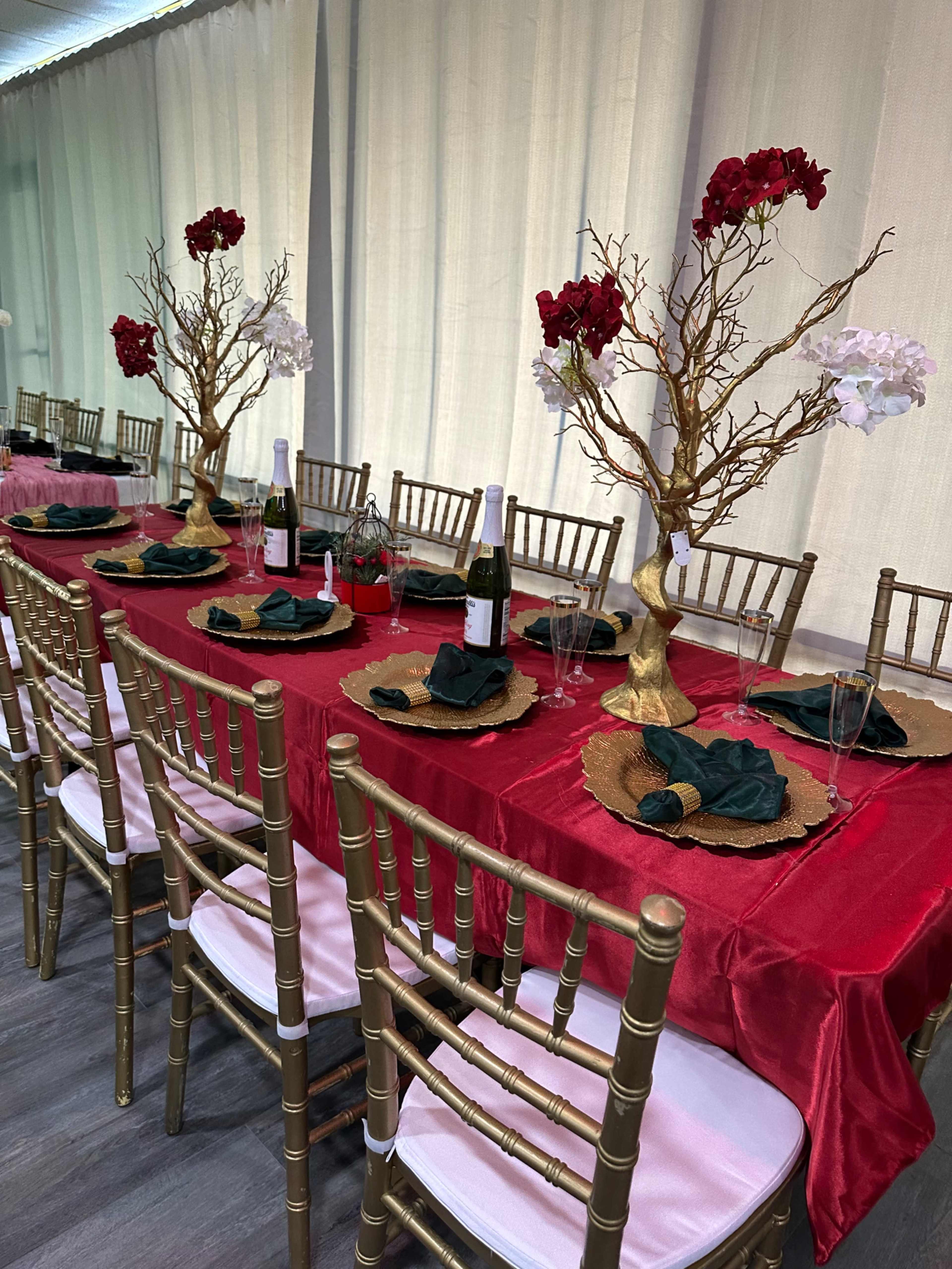 A long dining table is set with gold-charged plates, dark green napkins, and decorative centerpieces featuring branches with red and white flowers, draped in a red tablecloth.