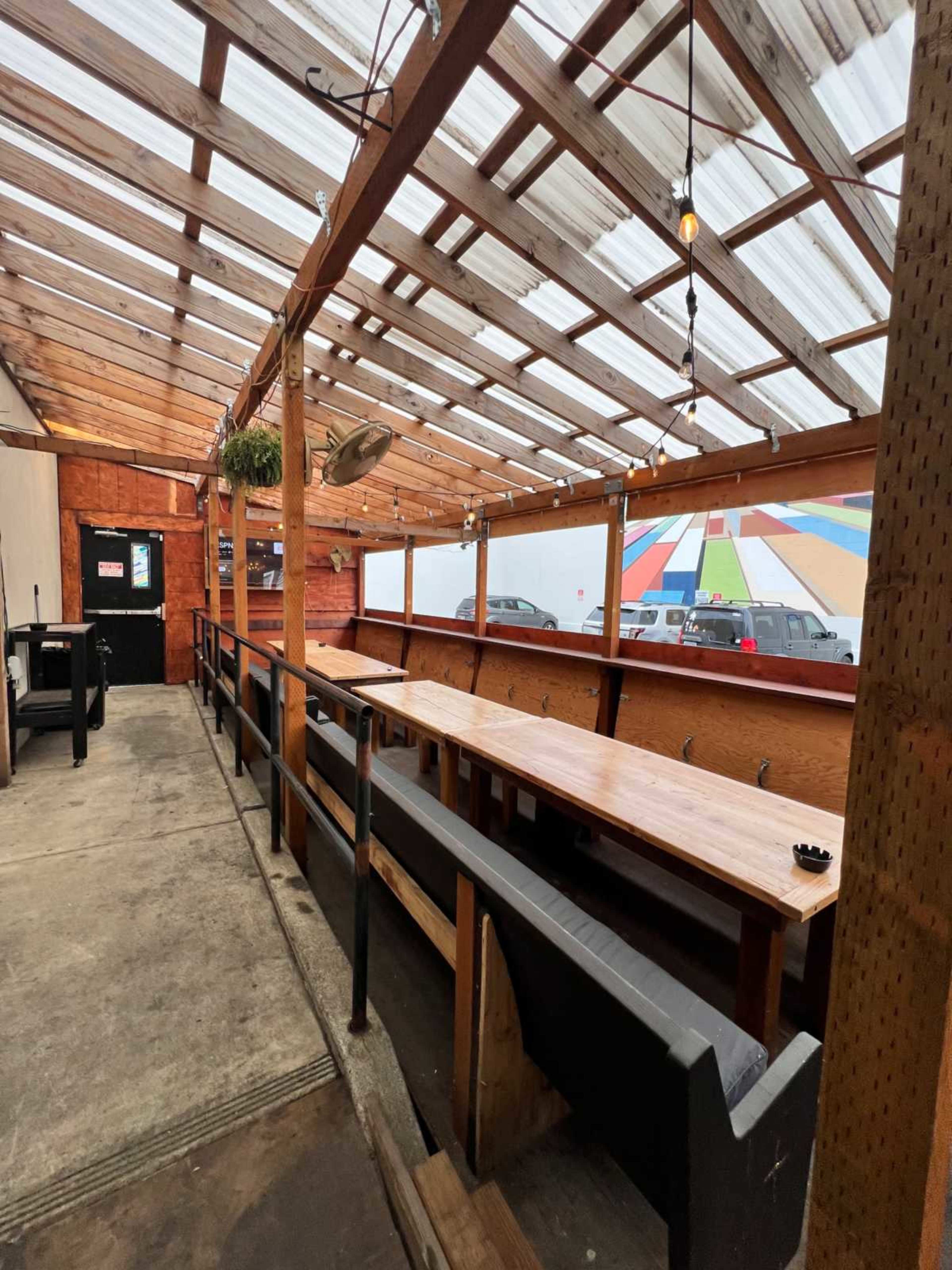 The image shows a spacious outdoor seating area with a wooden table and benches under a transparent roof, featuring string lights and a colorful mural in the background.