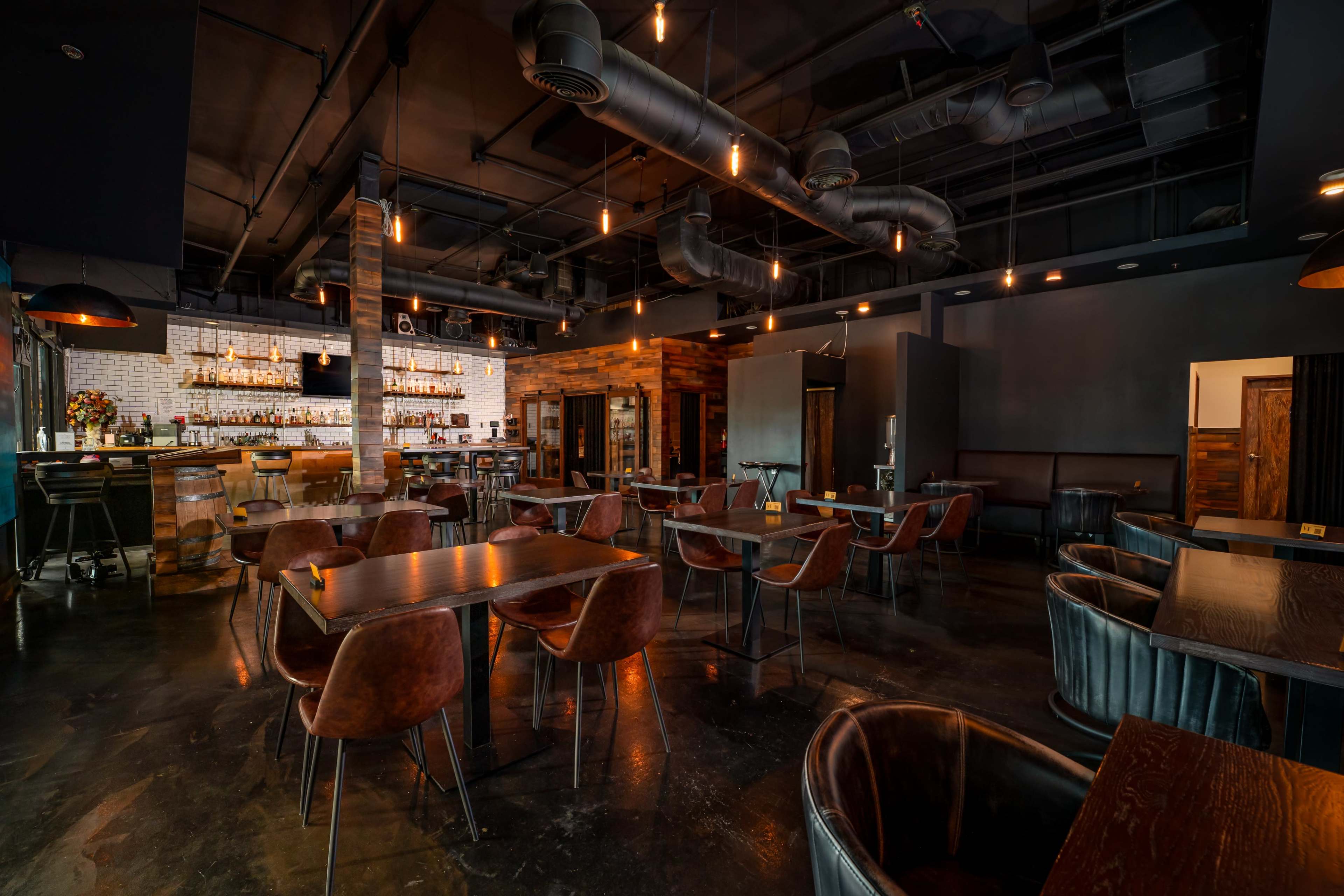 The image shows a modern restaurant interior featuring dark wood furniture, leather seating, and a well-lit bar area in the background.