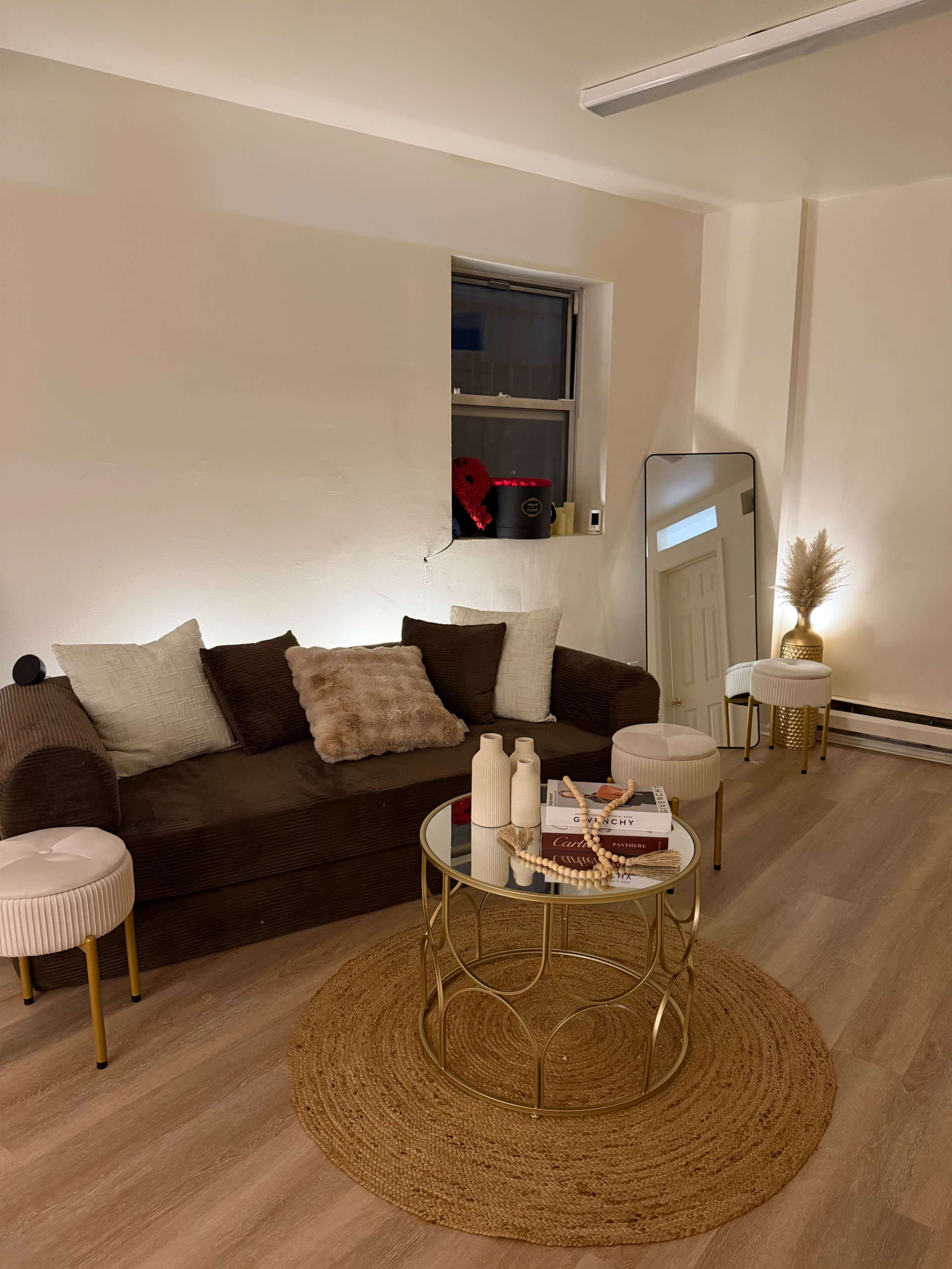 A cozy living room features a dark brown sofa with decorative pillows, a round glass coffee table atop a woven rug, and a large mirror next to light-colored decor elements.