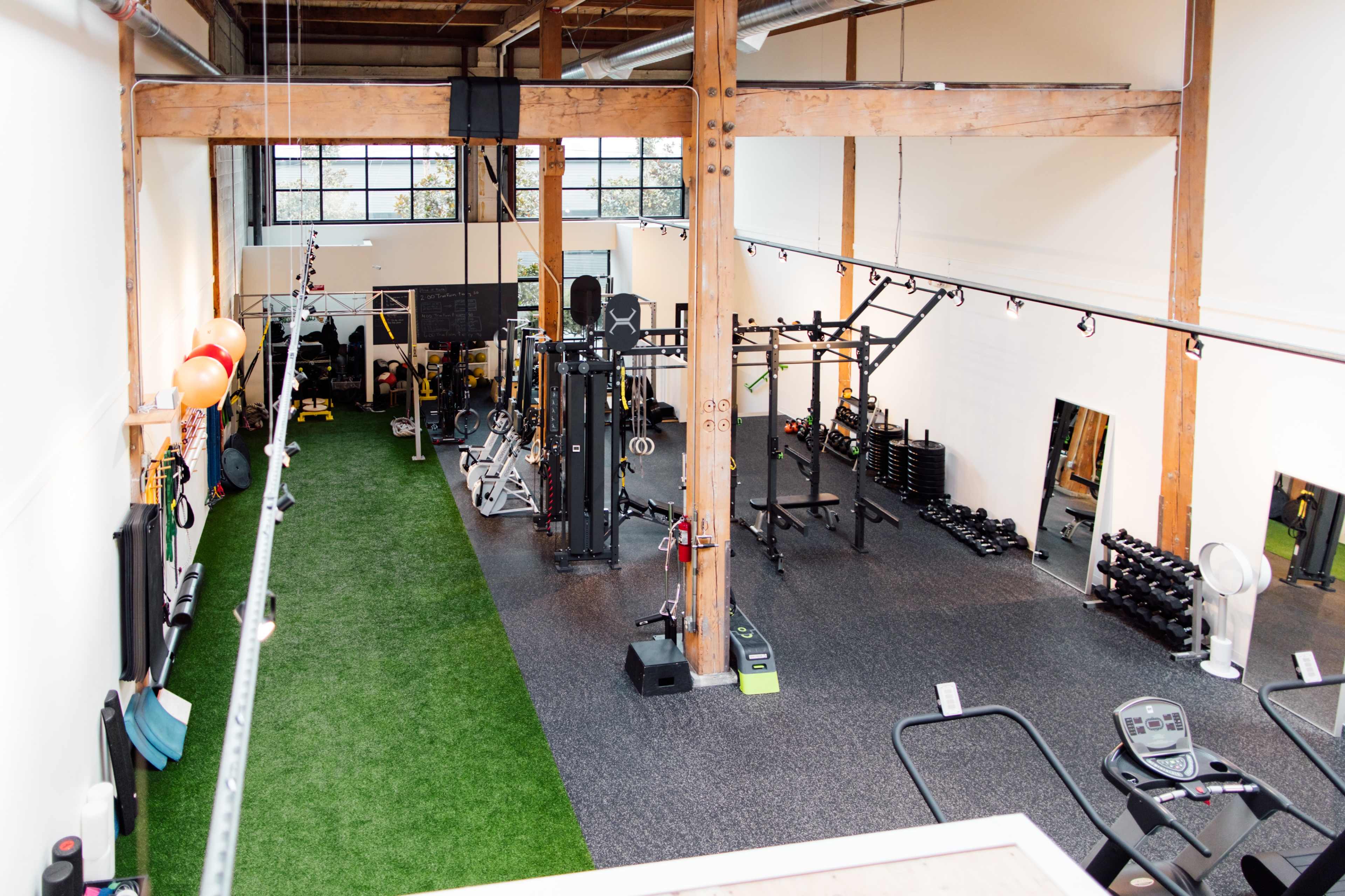 A spacious gym with various exercise equipment, including weights and cardio machines, situated on a carpeted floor with turf sections.