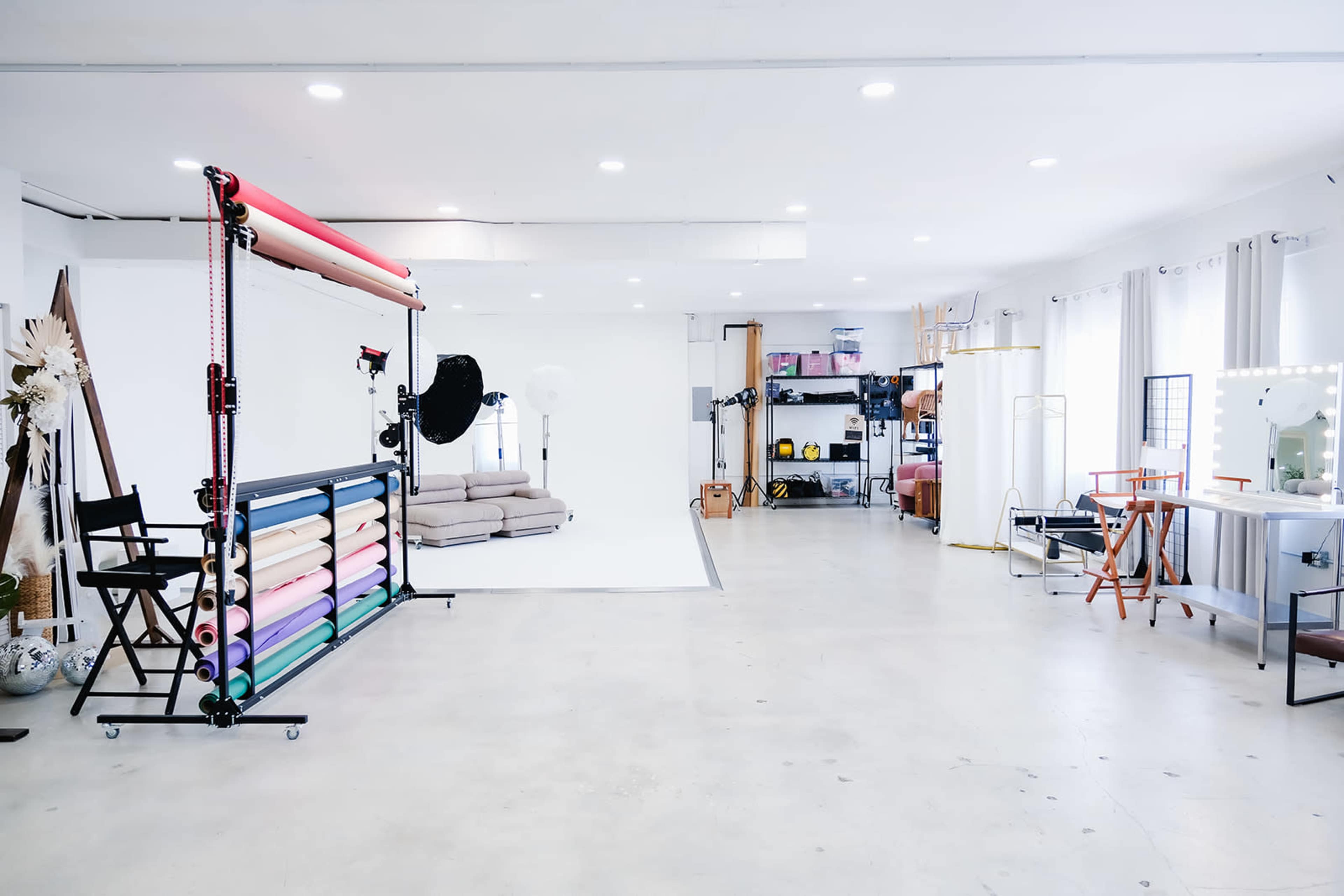 The image shows a photography studio with a clean, open space, featuring a backdrop setup, lighting equipment, and various props organized on shelves.