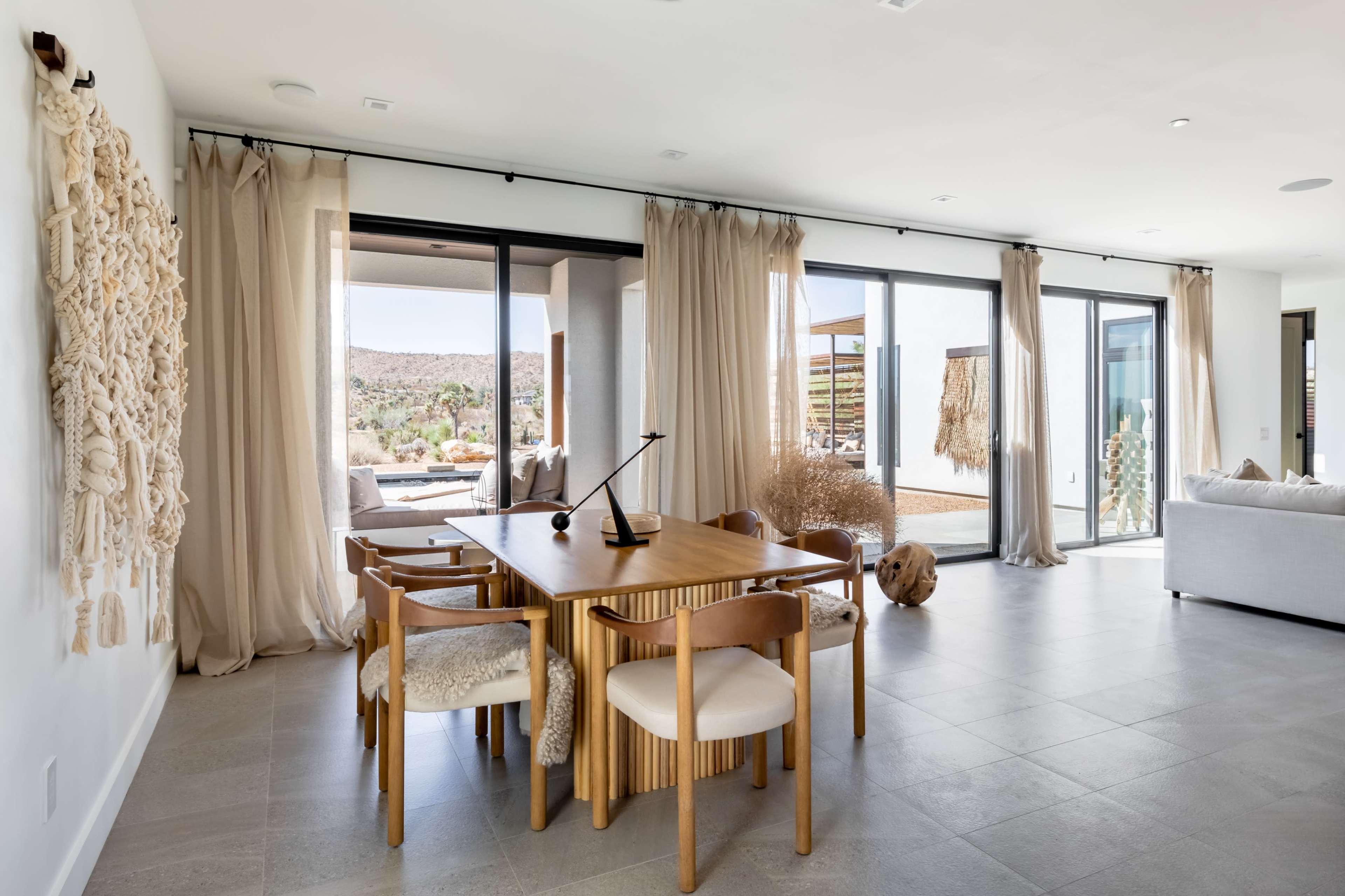 A modern interior features a wooden dining table surrounded by chairs, with large glass doors opening to a scenic outdoor view and soft drapes framing the space.