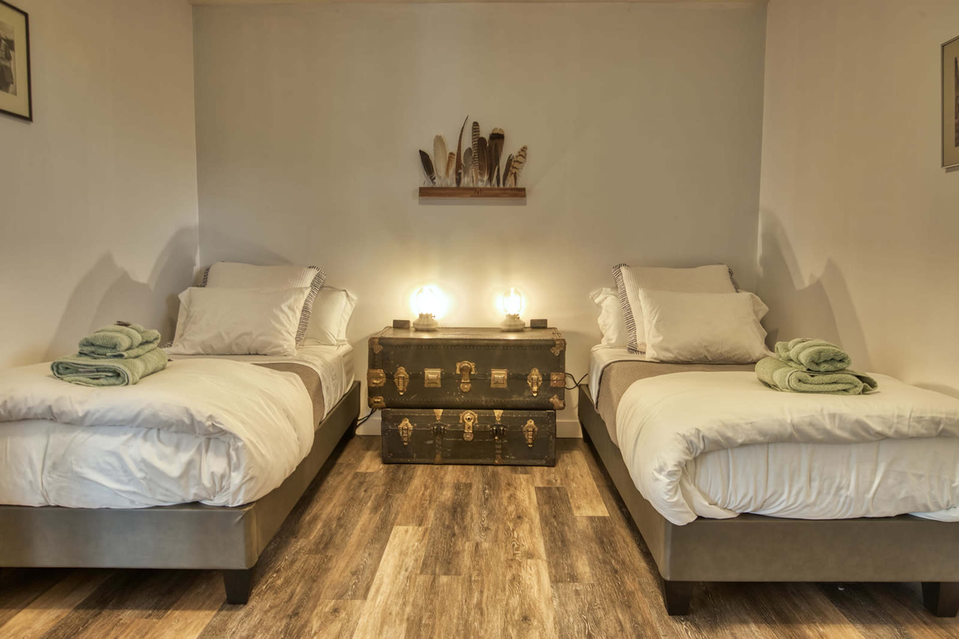 The image shows a cozy bedroom featuring two twin beds with white linens and green towels folded neatly on each bed, along with a vintage chest at the foot of the beds and a shelf with decorative feathers on the wall above.