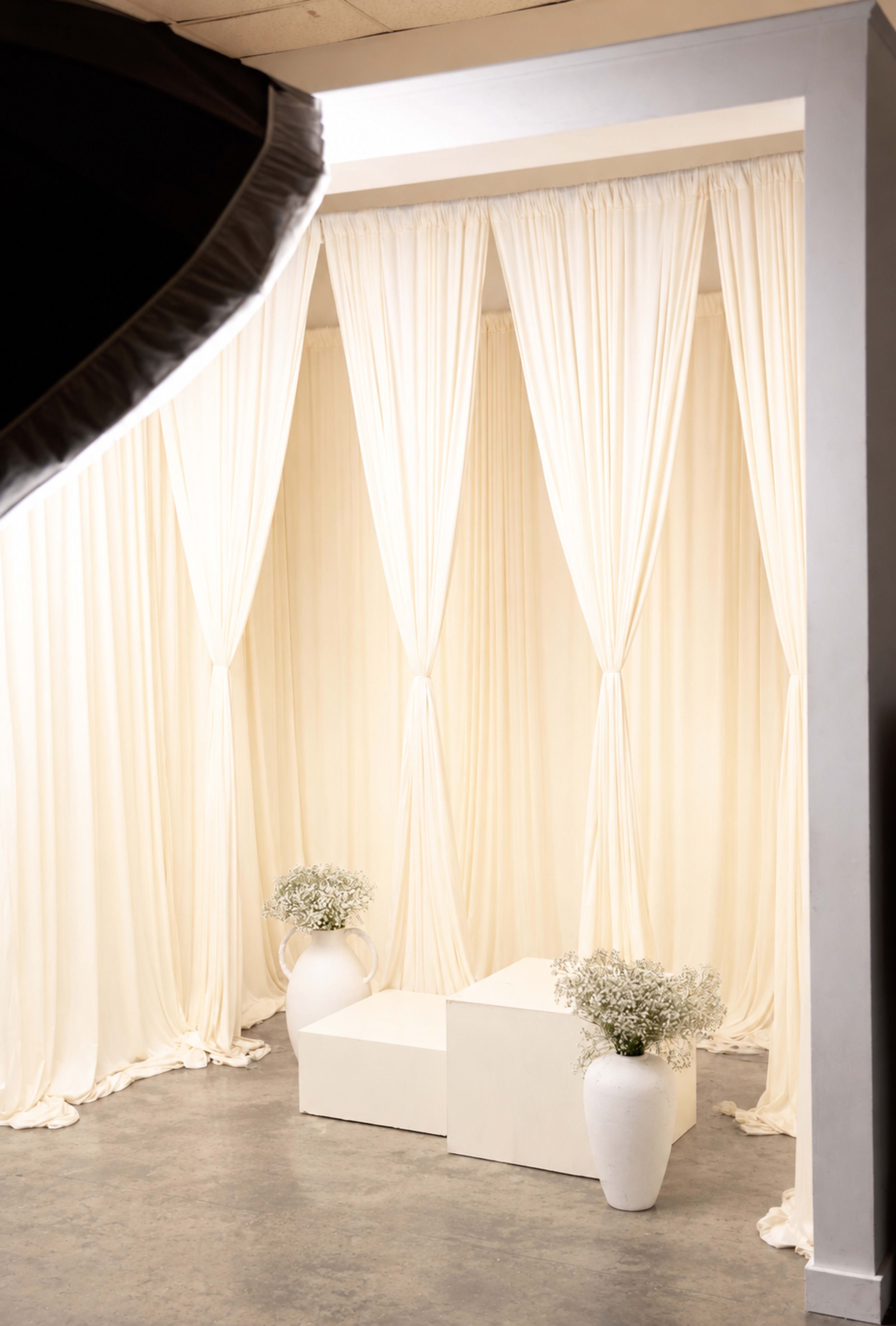 The image shows a minimalist studio setup with white draped curtains, two white vases with flowers, and a simple white platform.