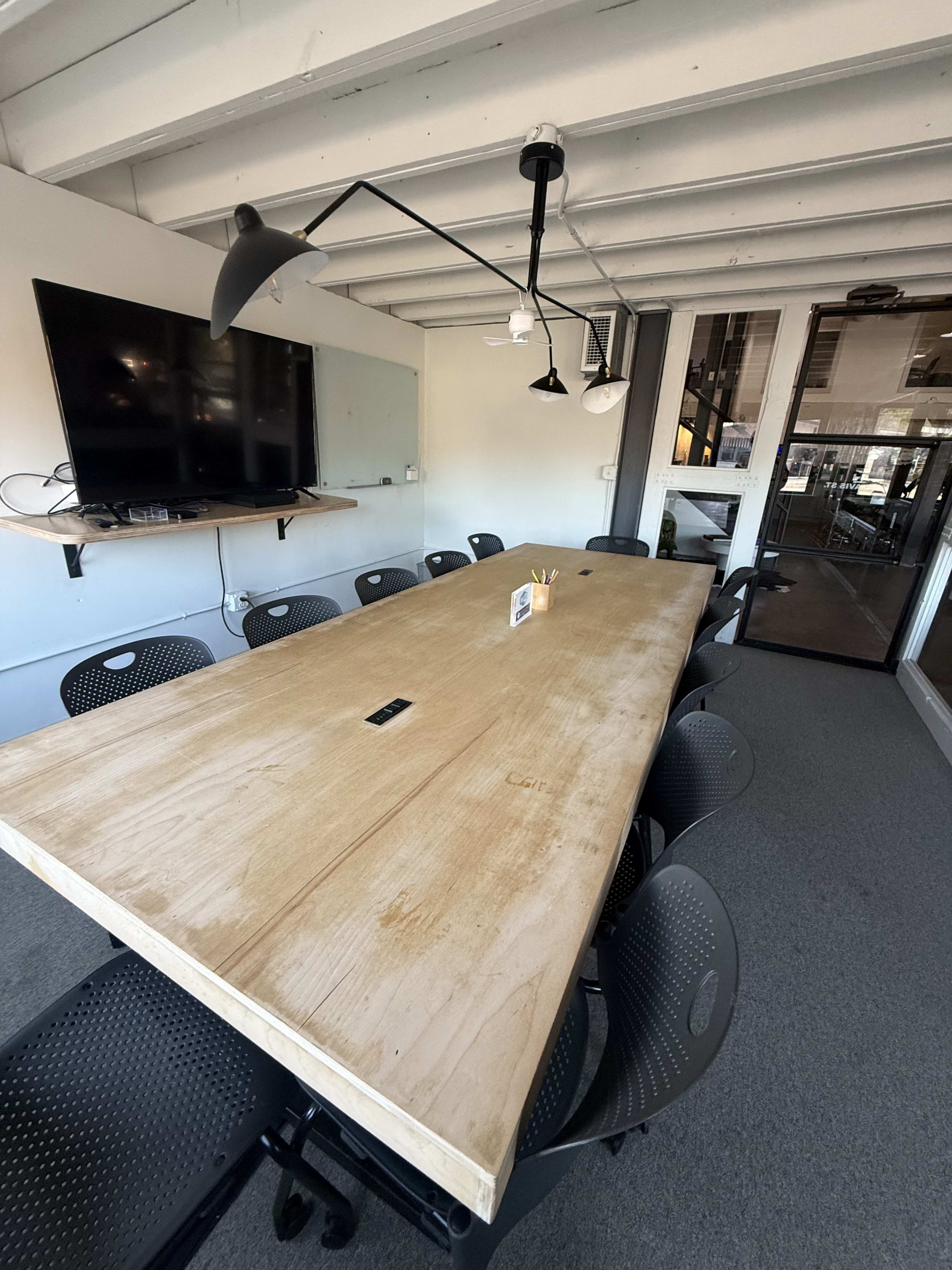The image shows a long wooden conference table surrounded by black chairs in a well-lit meeting room with a television mounted on the wall.