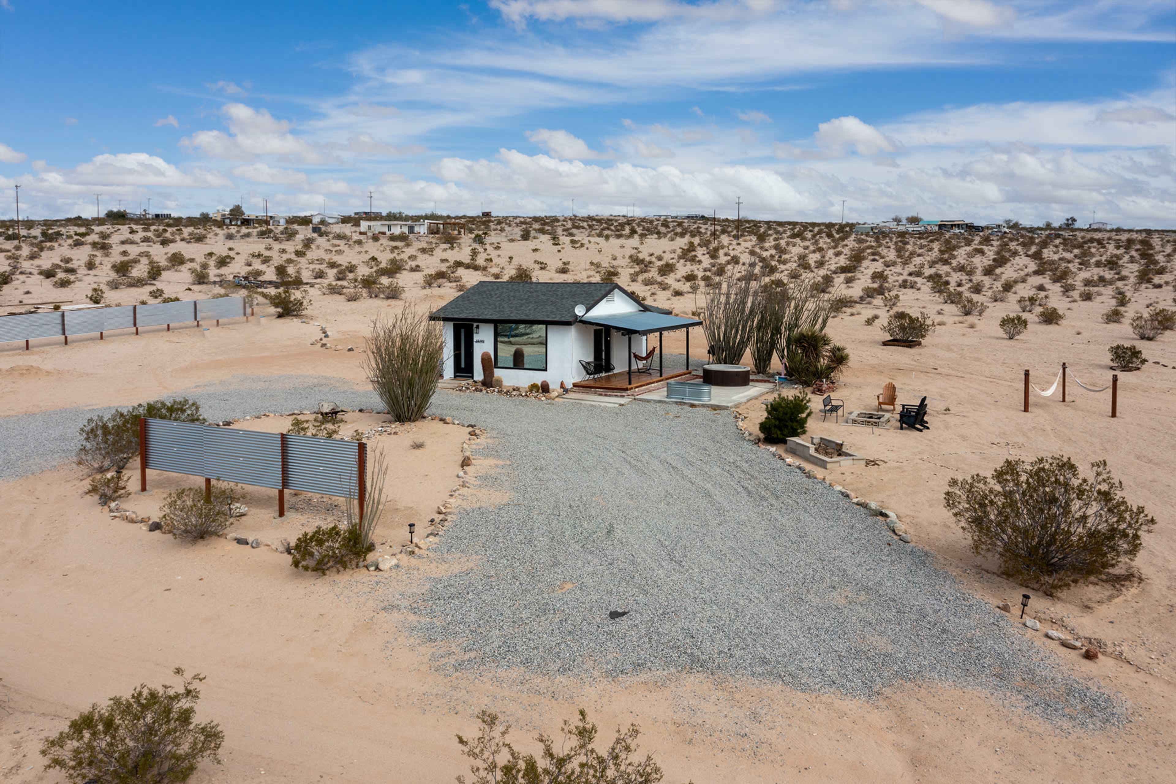 The image shows a small house surrounded by a gravel driveway and sparse desert vegetation, set against a backdrop of a blue sky with scattered clouds.