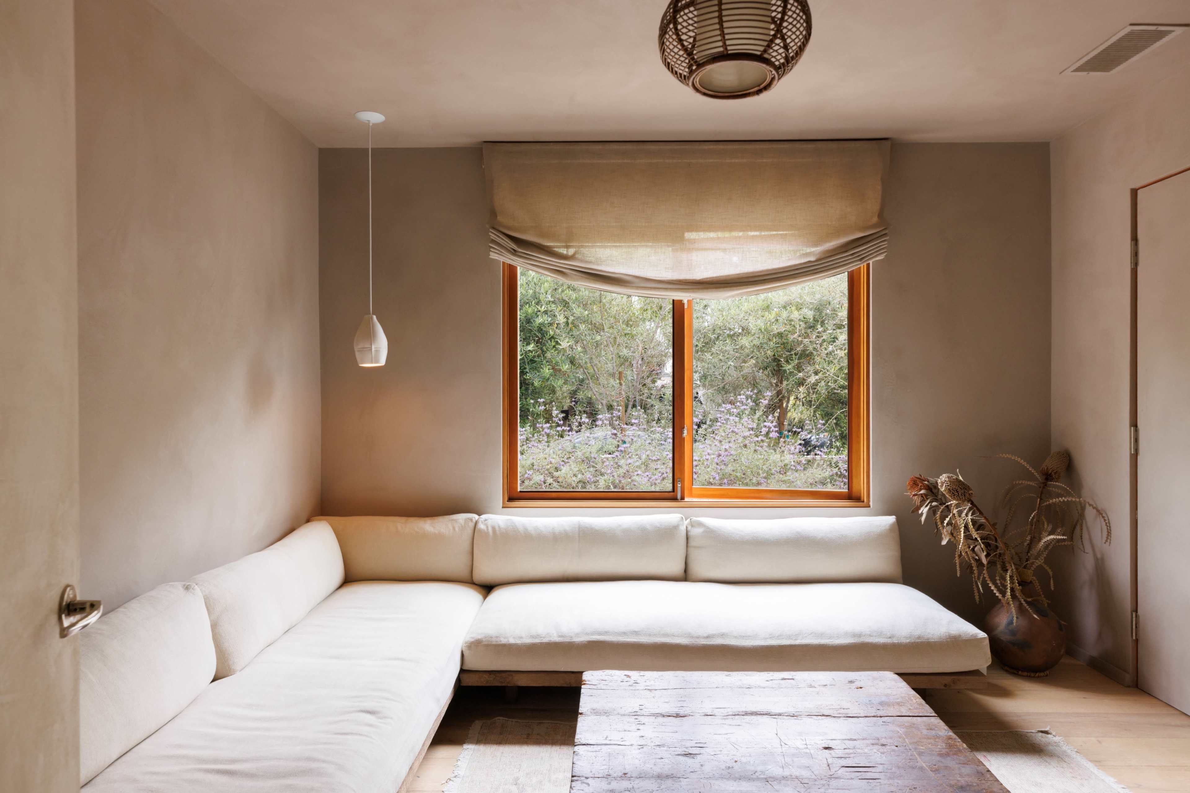 A minimalist living room features a L-shaped white couch, a wooden coffee table, and a large window overlooking greenery.