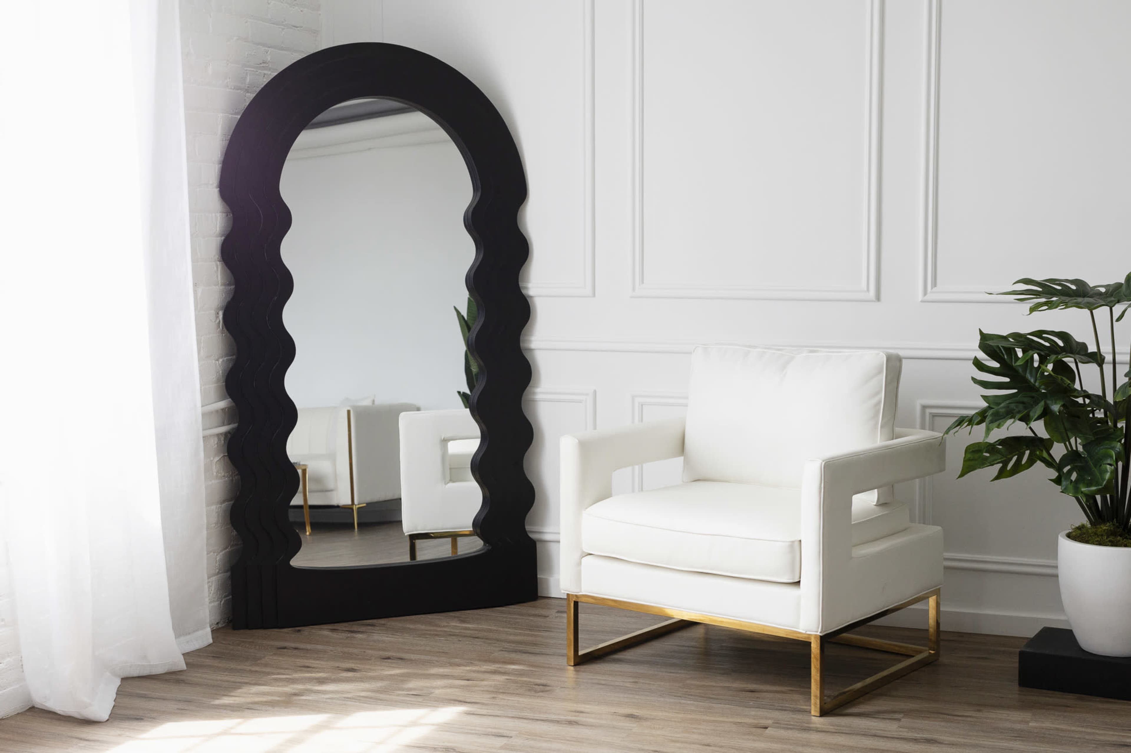 A large black-framed mirror stands next to a white armchair with gold legs in a bright, minimalist room.