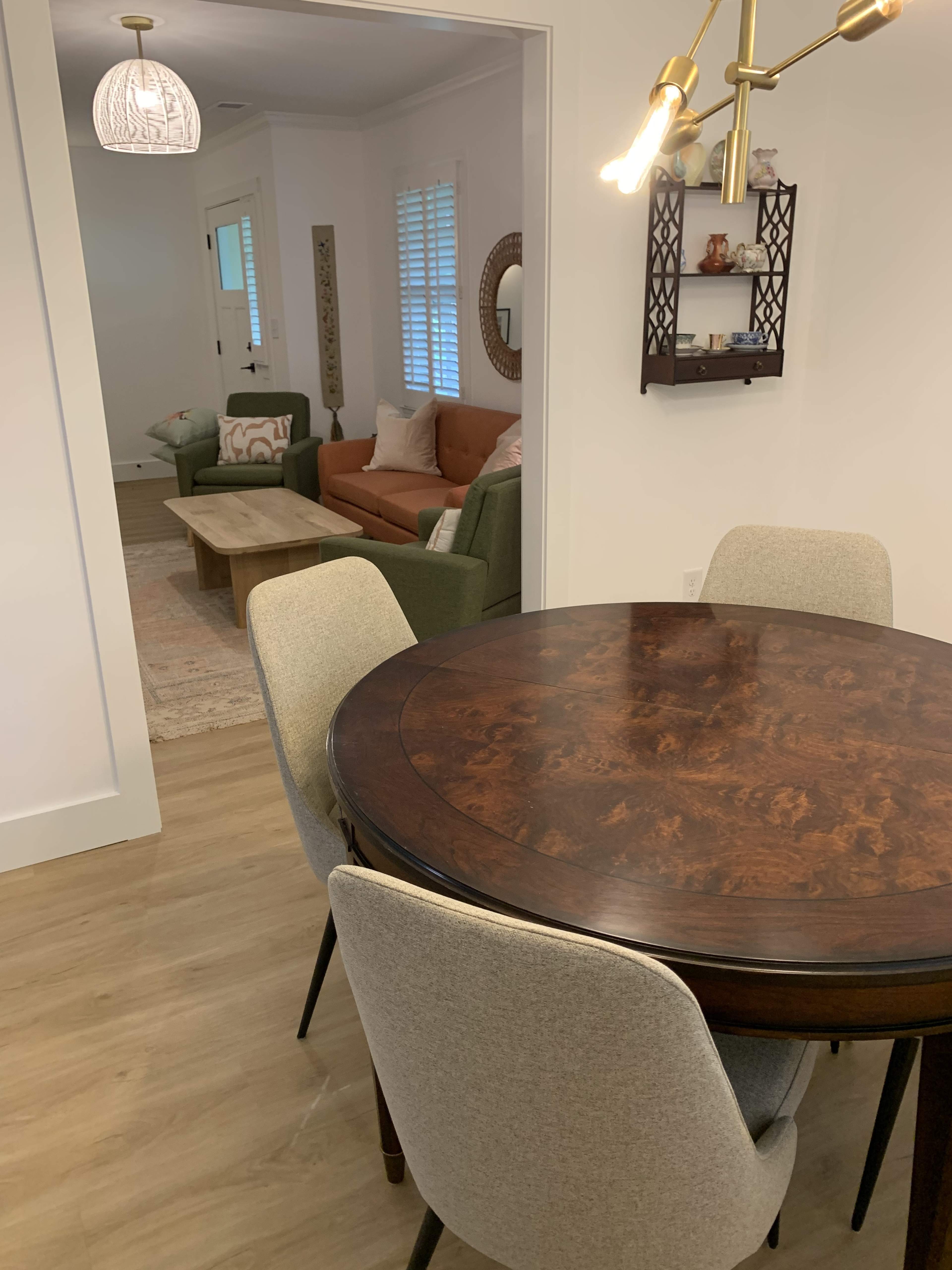 A round wooden dining table with four upholstered chairs is placed in a bright, open room that leads to a cozy living area with a green sofa and a wooden coffee table.