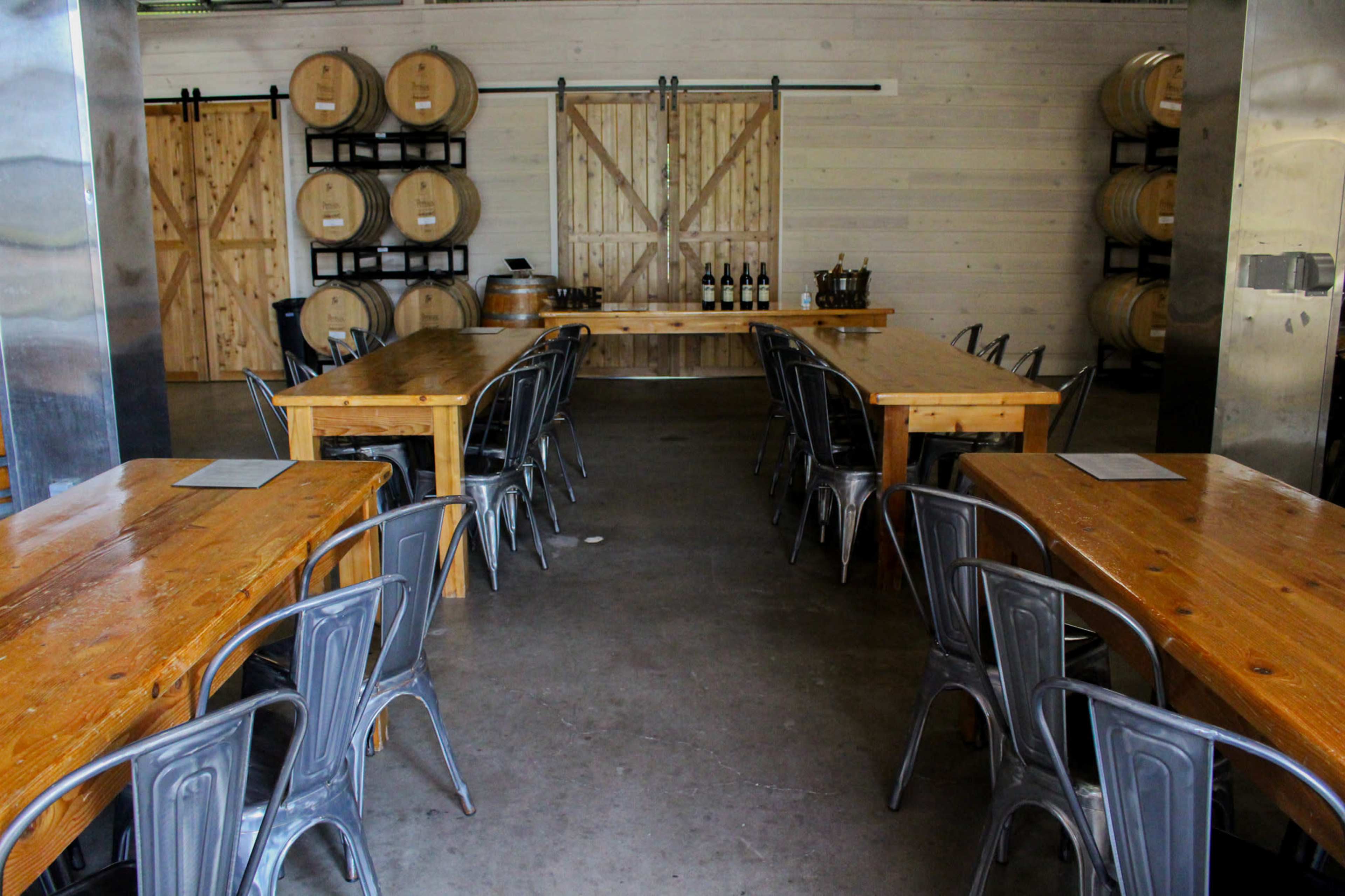 A spacious tasting room features several wooden tables lined with metal chairs, barrels stored against the walls, and a setup for wine display at the back.