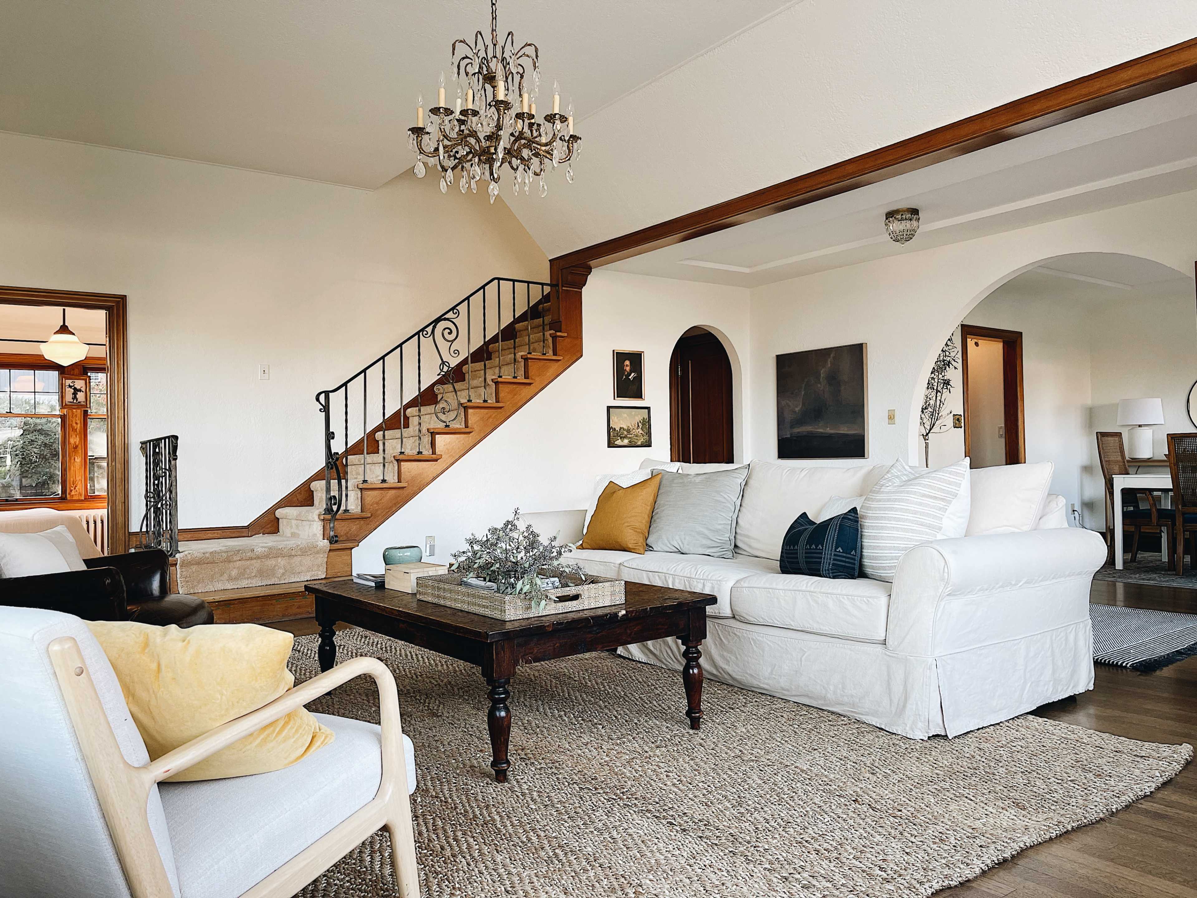 A living room with a white sofa, a dark wooden coffee table, a staircase in the background, and a chandelier hanging from the ceiling.